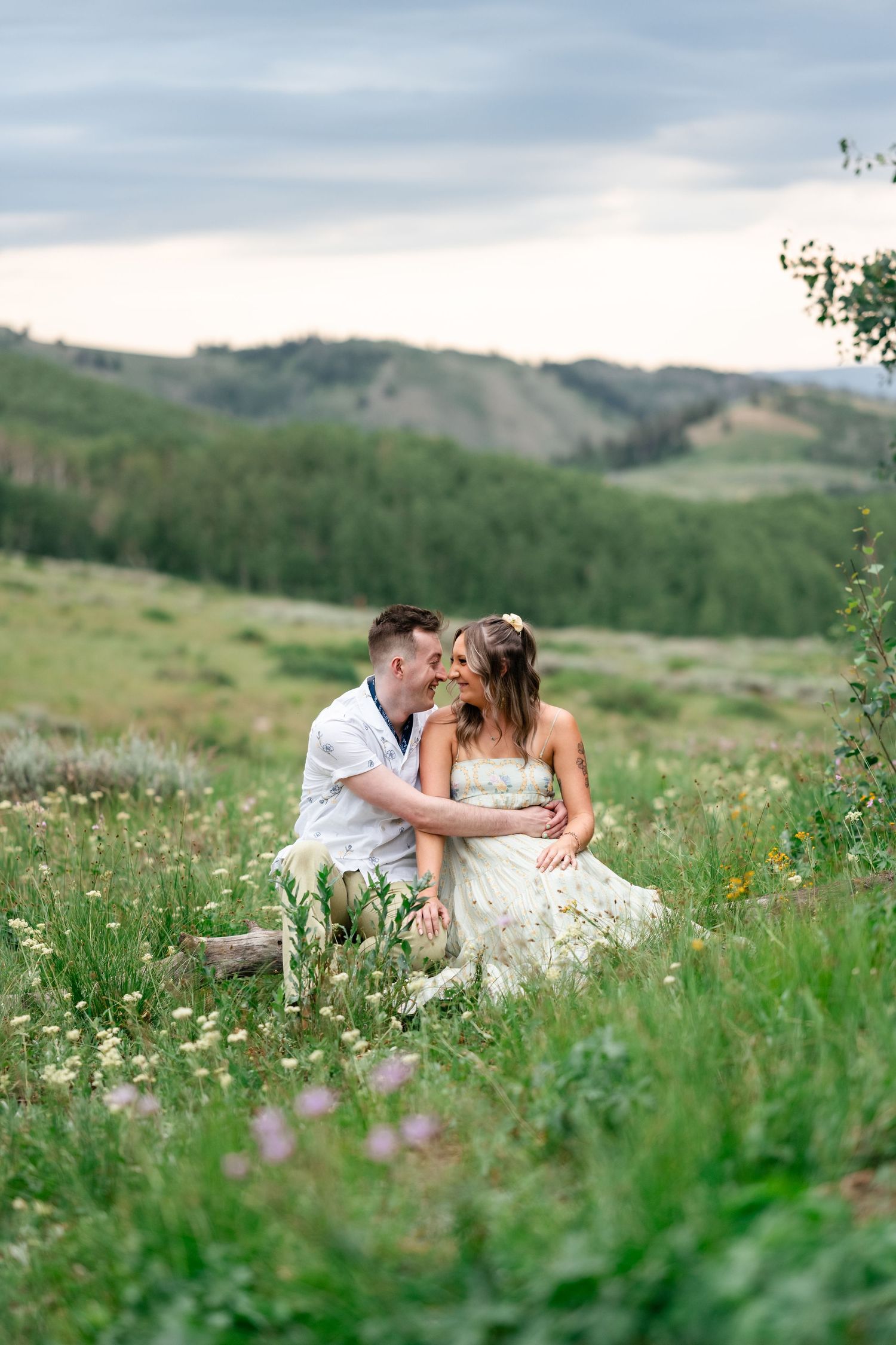 A couple sits intimately in a wildflower meadow with rolling hills and mountains in the background.