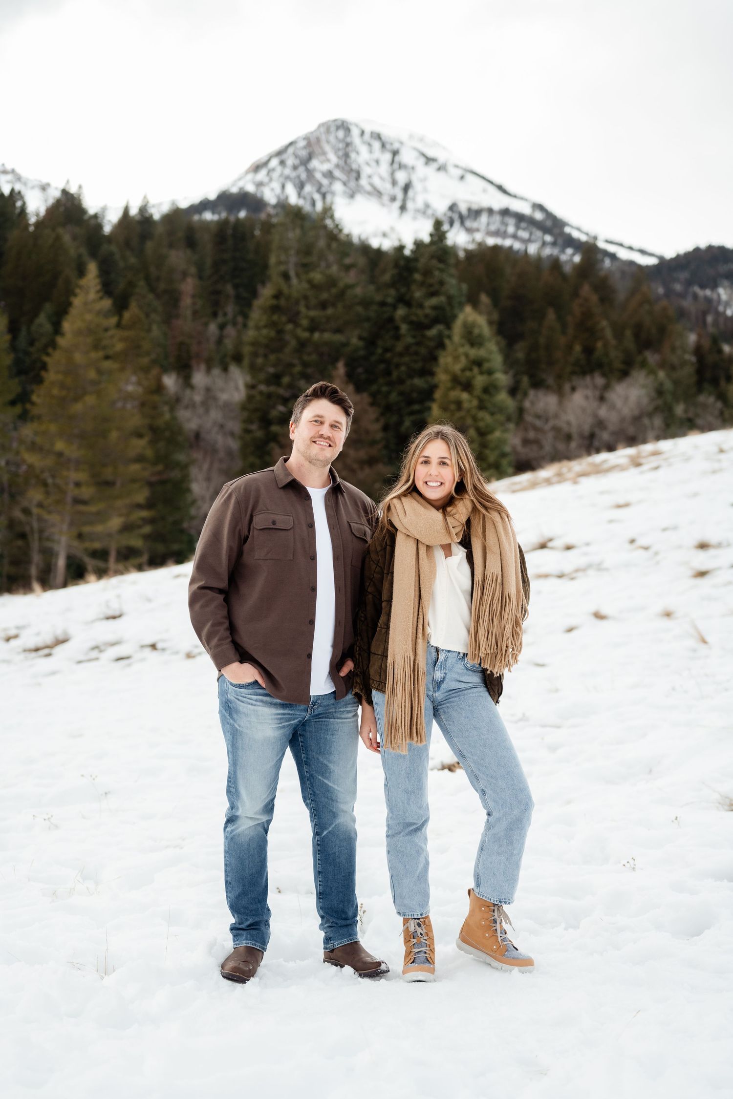 A couple poses together in a snowy winter landscape with mountains and evergreen trees in the background.