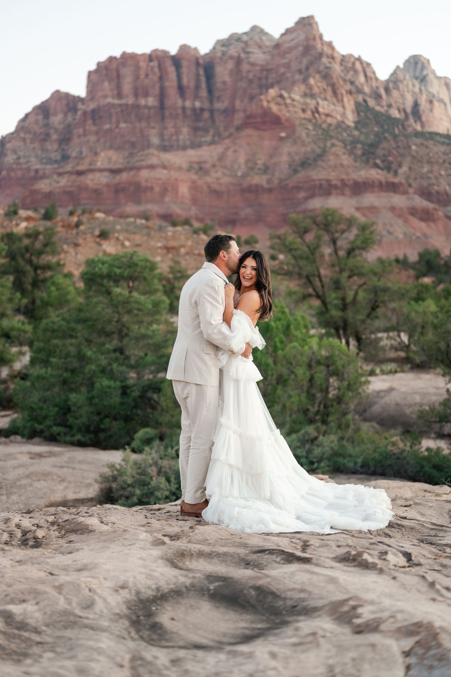 Zion destination photographer. A romantic wedding couple embraces on a rocky ledge with majestic red rock cliffs in the background.