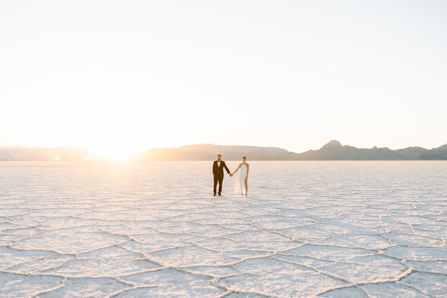 Two silhouettes hold hands while standing on the vast white expanse of the Bonneville Salt Flats at sunset. Salt Flats Photographer.