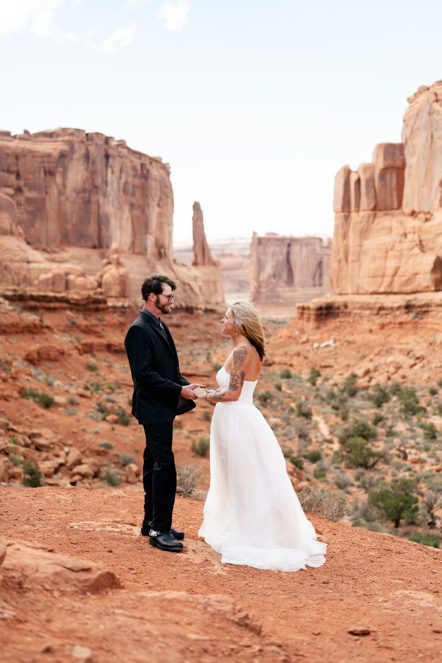 A couple in wedding attire stands on red desert cliffs at Arches National Park with dramatic rock formations in background.
