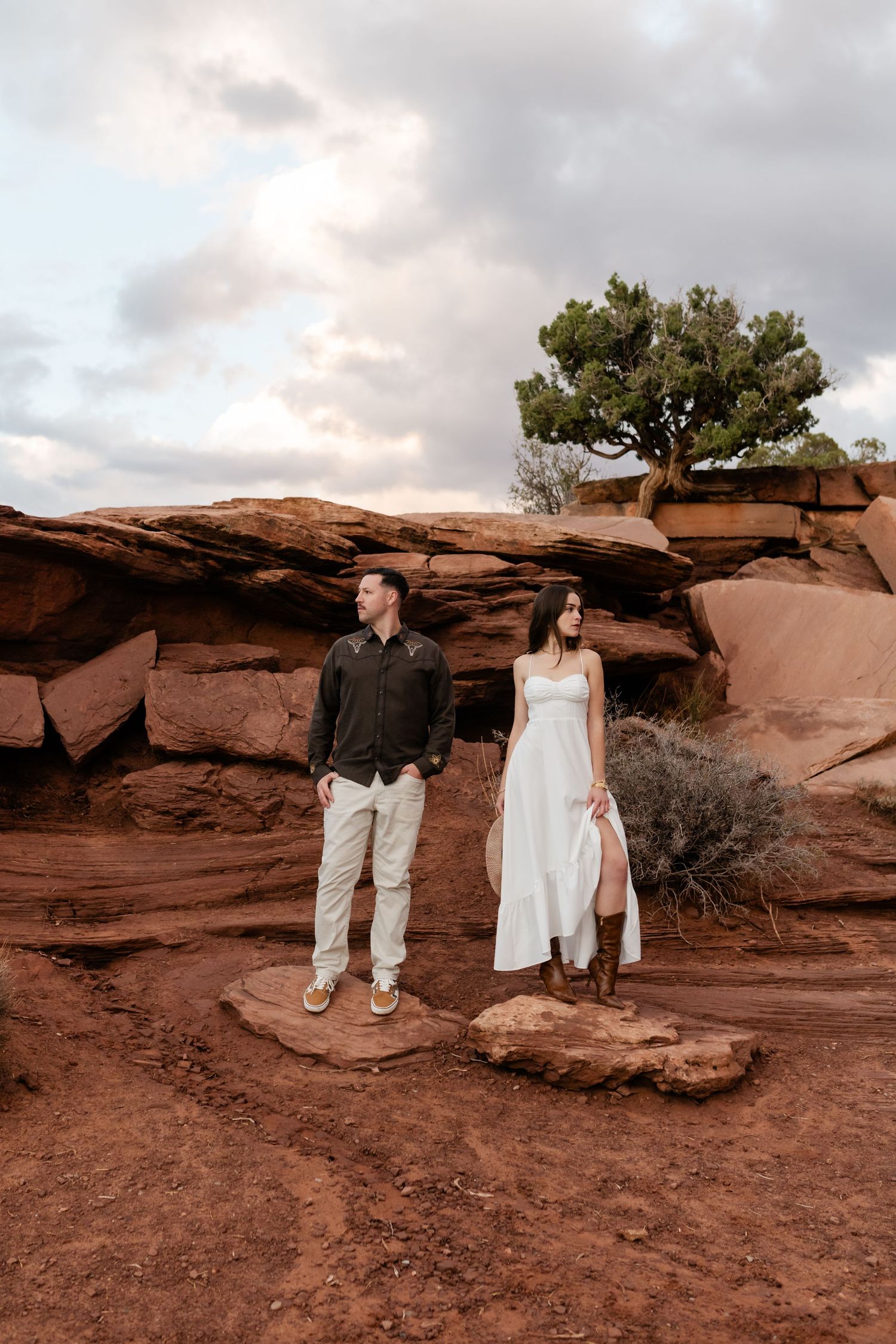 A couple poses on red rock formations with a lone tree and cloudy sky backdrop.