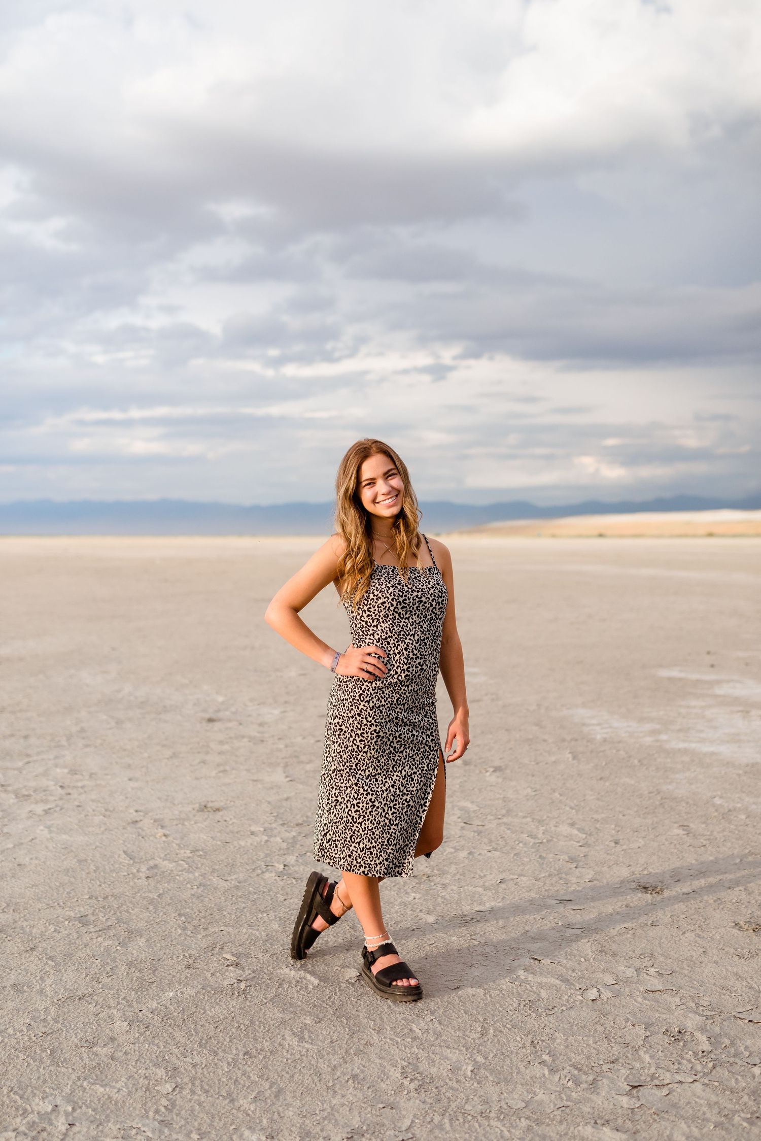 A person in a leopard print dress and sandals poses on a vast salt flat under dramatic cloudy skies.