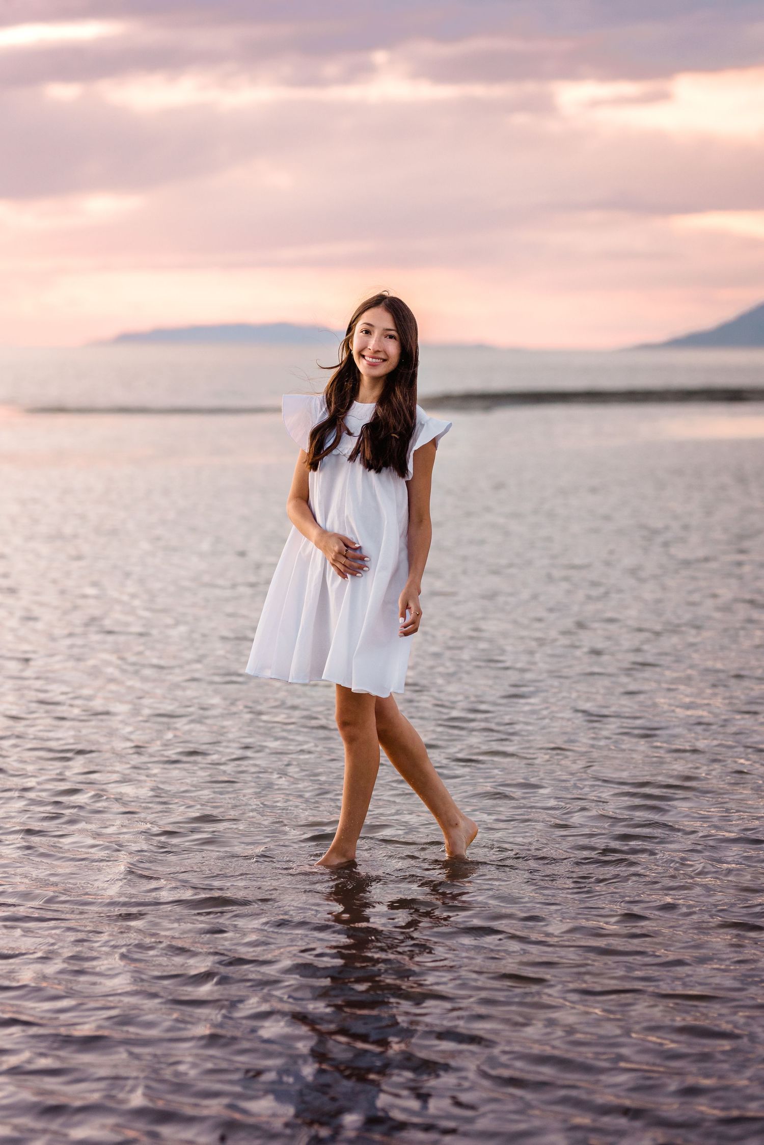 Person in flowing white dress wades through shallow water during golden hour at the beach.