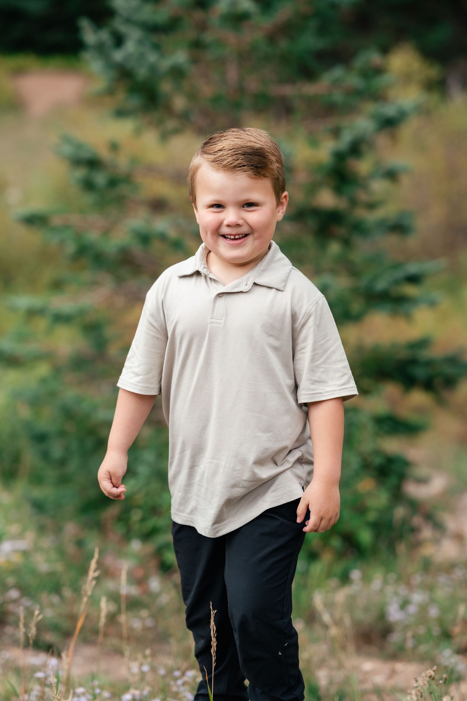 Young child in beige polo shirt smiling outdoors with evergreen trees in background.
