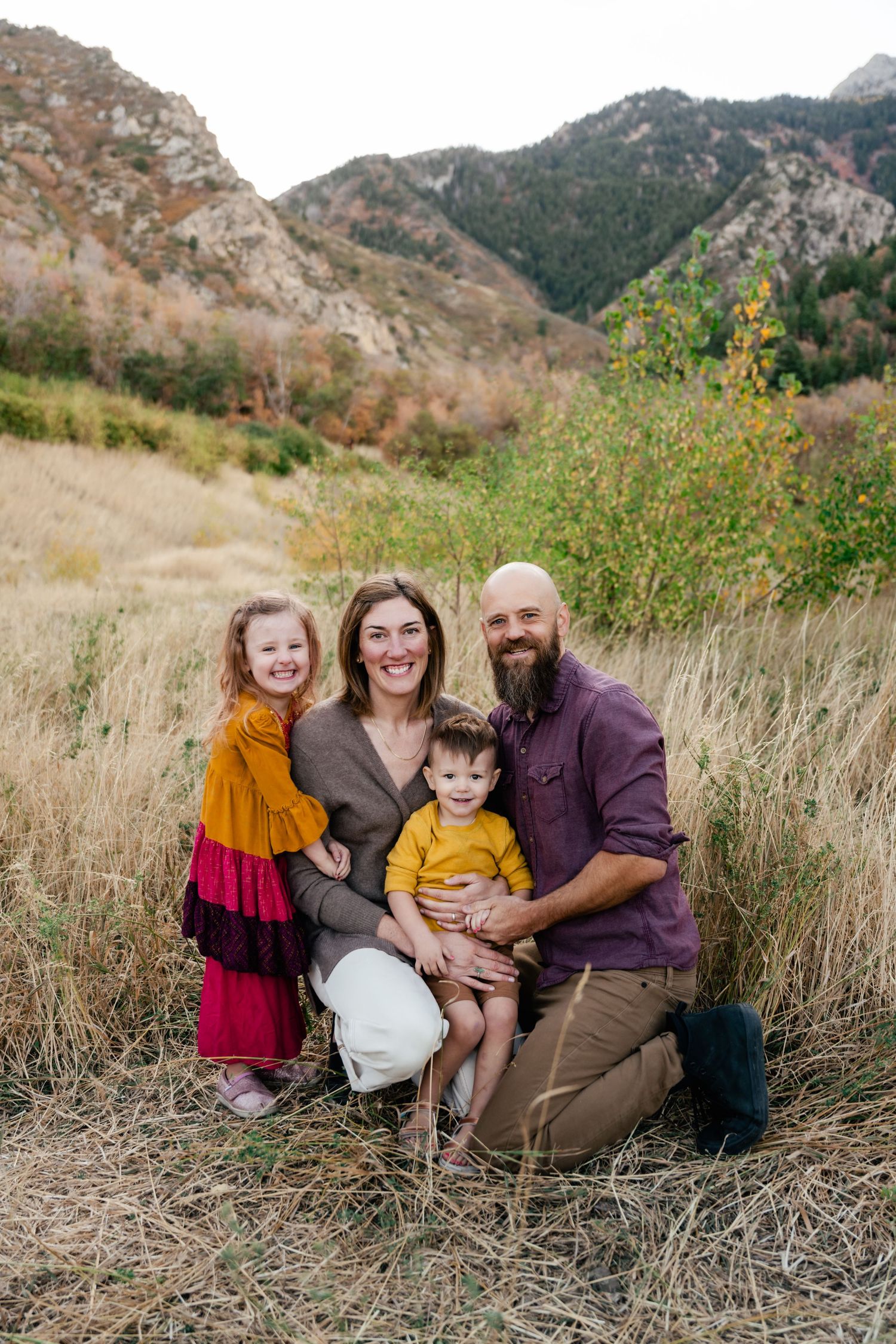 A family sits together in tall grass with mountains in the background during an autumn photoshoot.