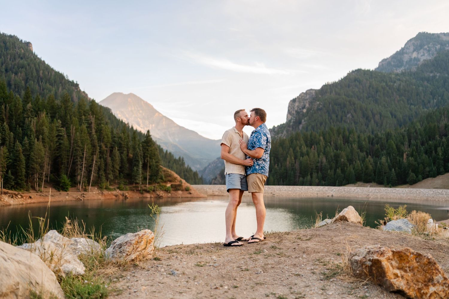 LGBTQ friendly photographer in Utah. Couple kissing by a mountain lake surrounded by pine trees and rocky terrain at sunset.