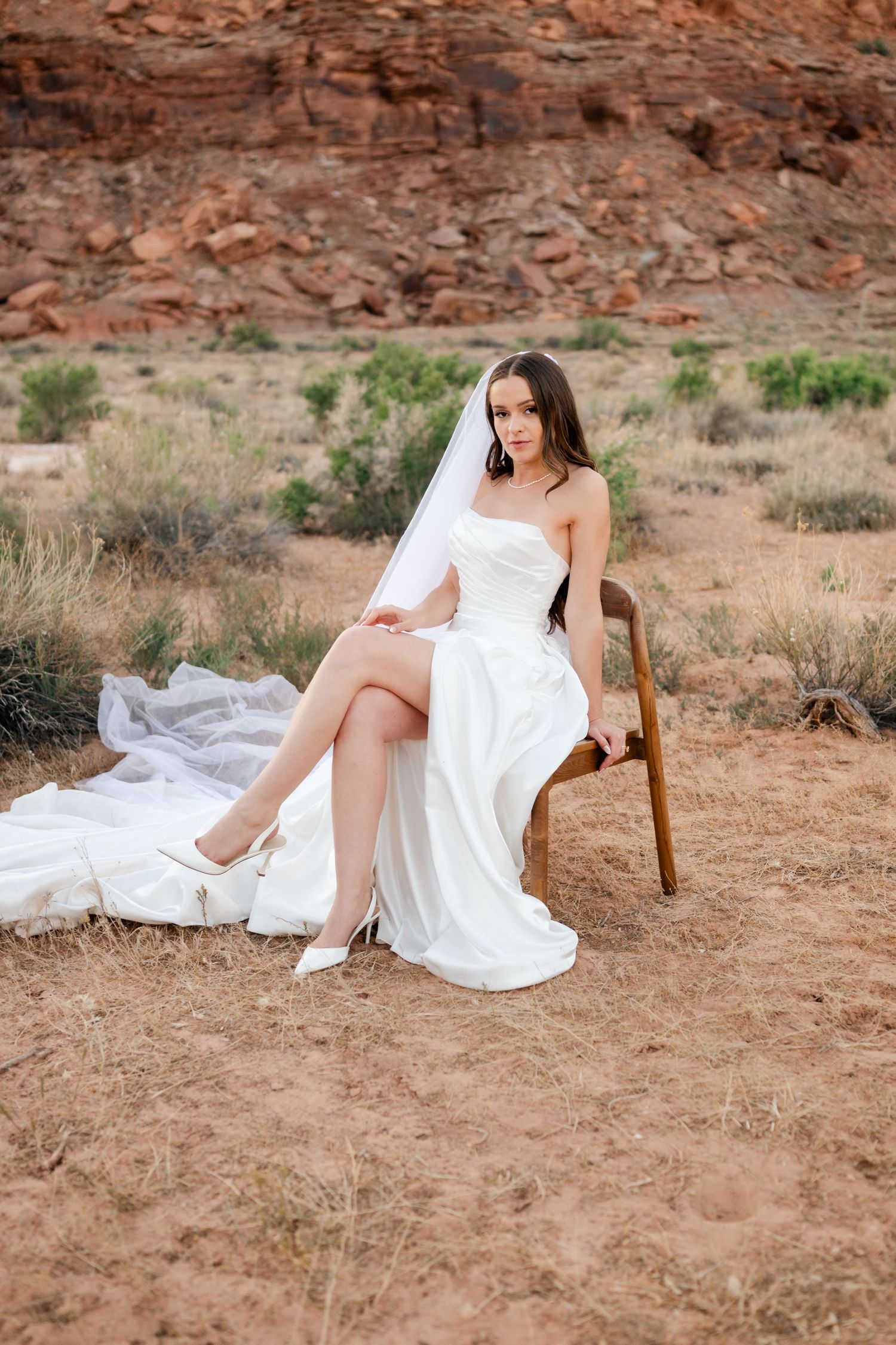 Bride in white strapless gown with veil sits on chair in red rock desert setting.