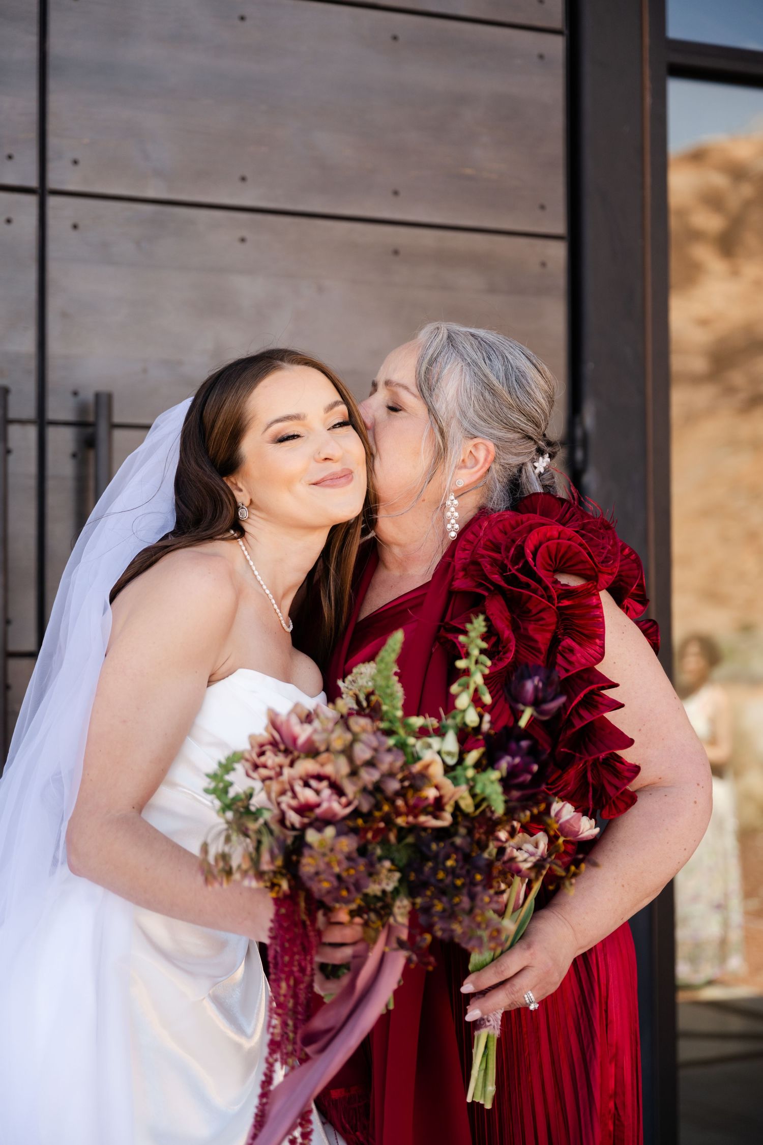 A bride in a white gown and veil receives a kiss from an older woman in a red dress, both holding bouquets.