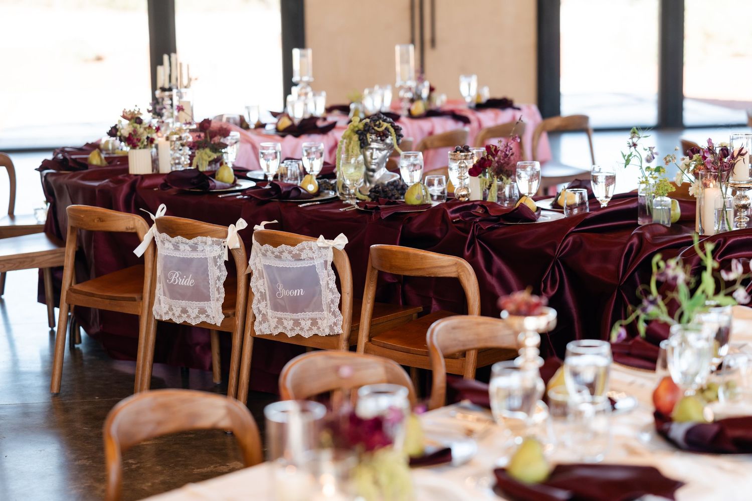 Elegant wedding reception with burgundy velvet tablecloths, wooden chairs, floral centerpieces, and lace chair signs in a bright venue.