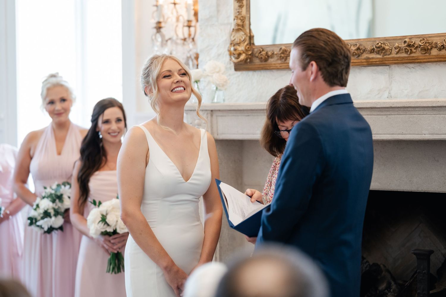 Bride smiling joyfully during wedding ceremony with bridesmaids in pink dresses holding white bouquets beside elegant fireplace.