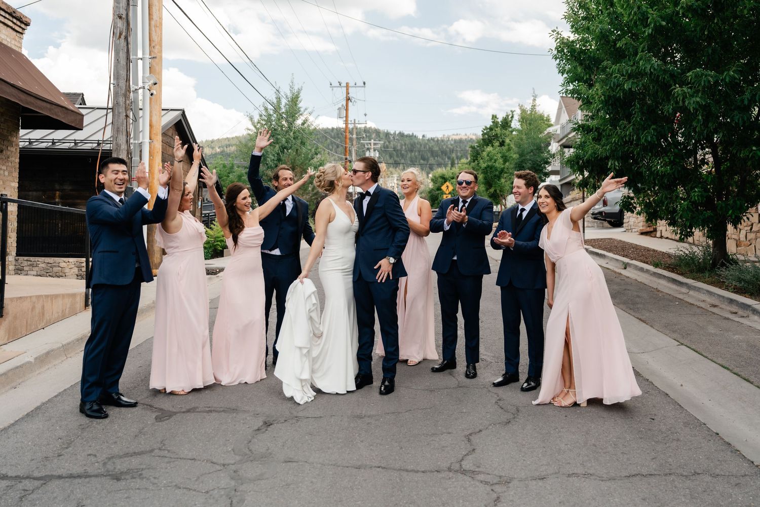 Wedding party in navy suits and blush gowns celebrating on a Park City street with mountains in the background.