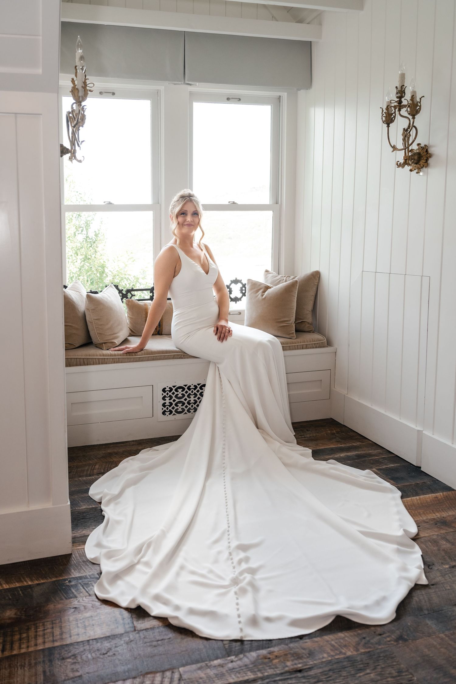 Bride in elegant white mermaid gown with long train sits on window bench at her intimate Park City Wedding.