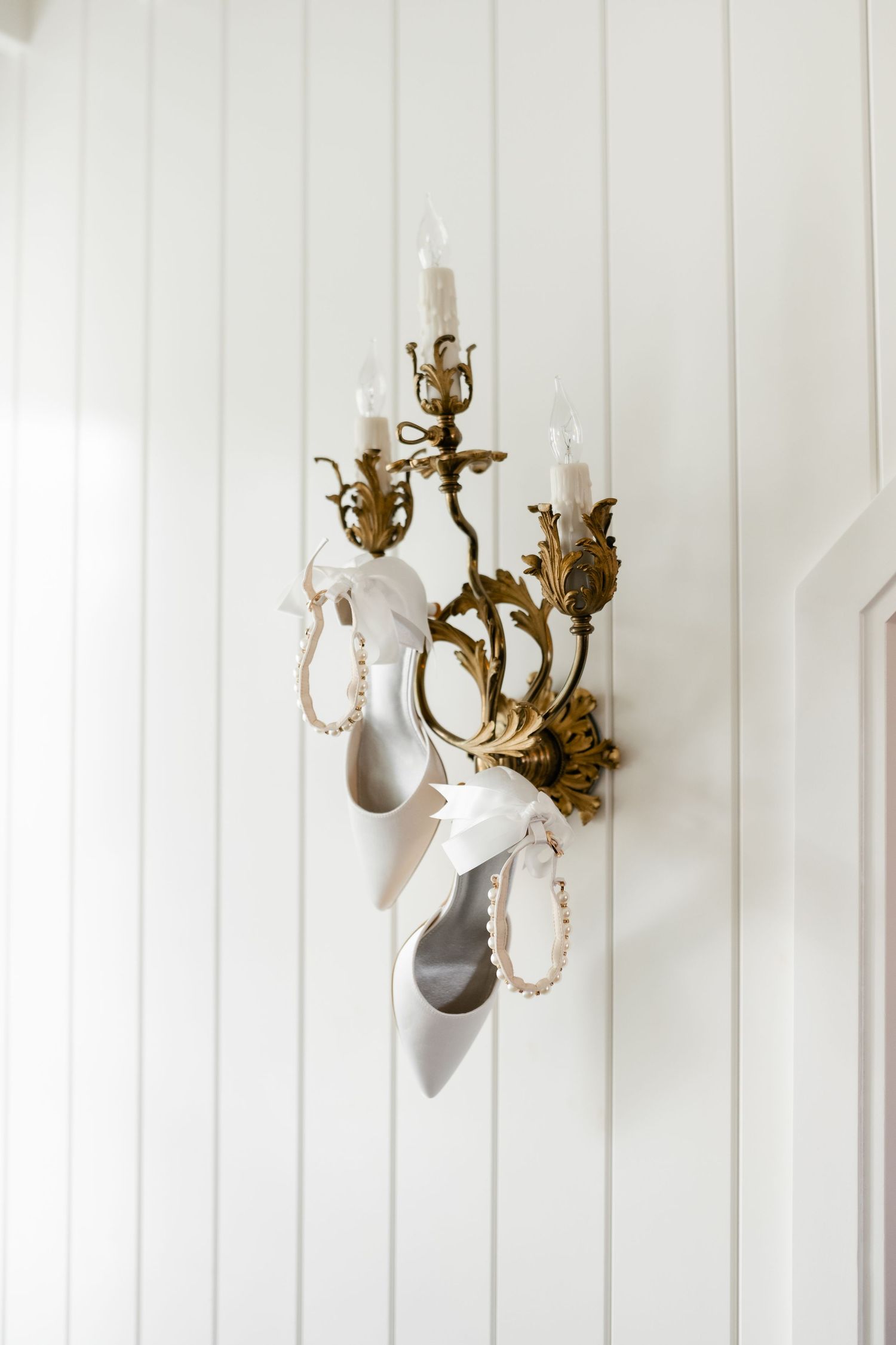 White bridal heels with pearl ankle straps hanging on an ornate gold candelabra wall sconce against white paneling.