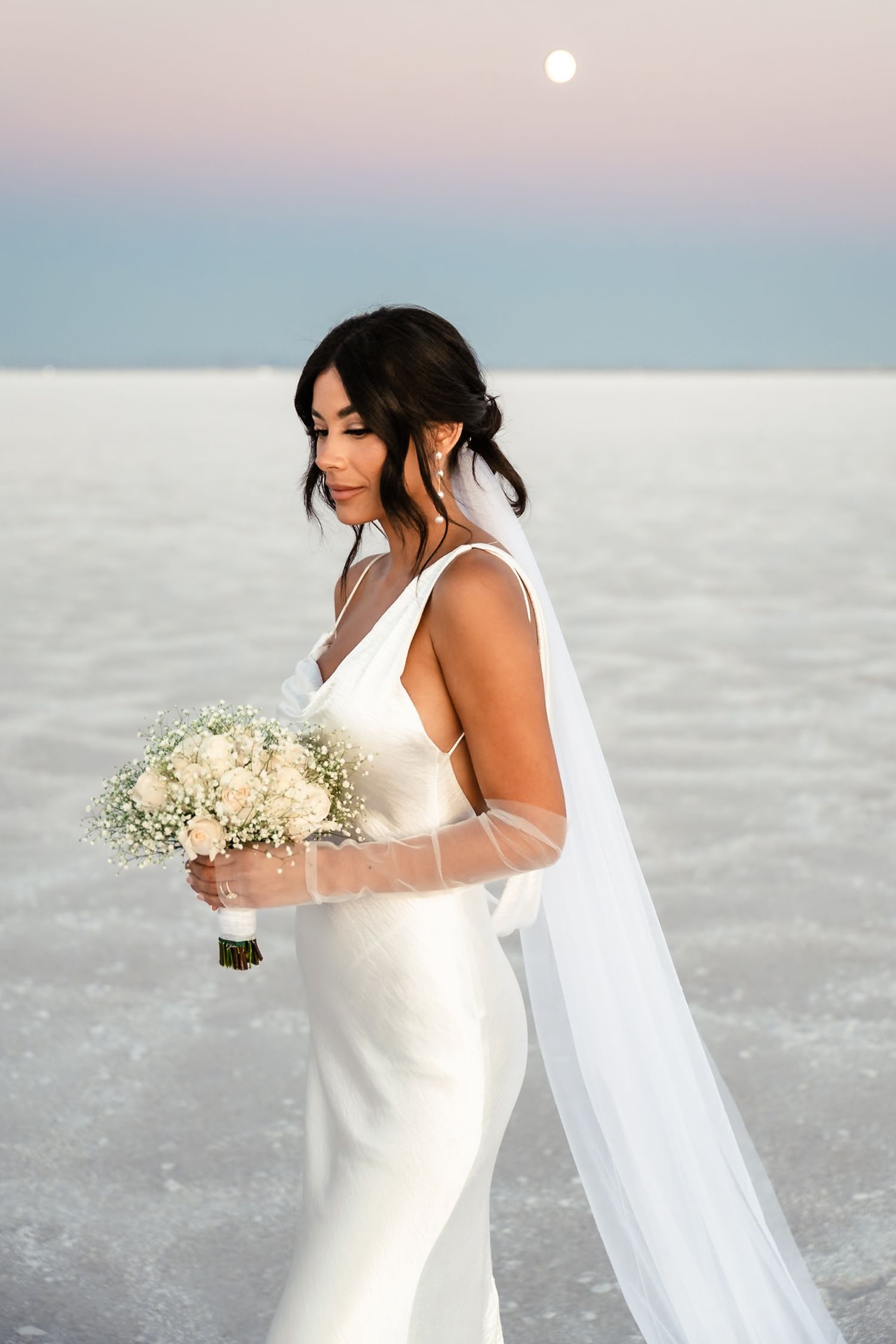 A bride holds a white bouquet against a pastel sunset sky on a salt flat.