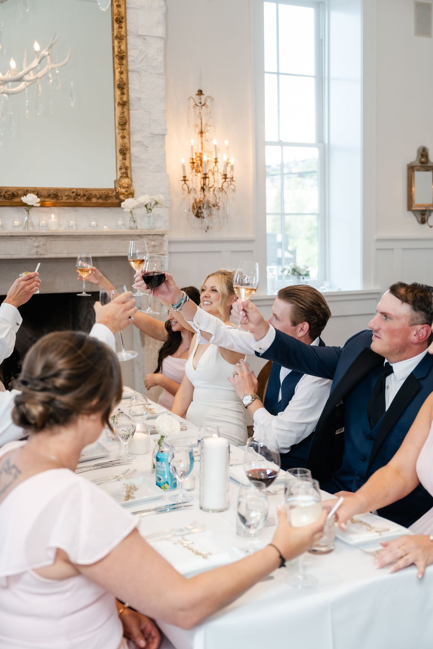 Wedding guests raise champagne glasses in an elegant white dining room with crystal chandeliers and ornate gold mirrors.
