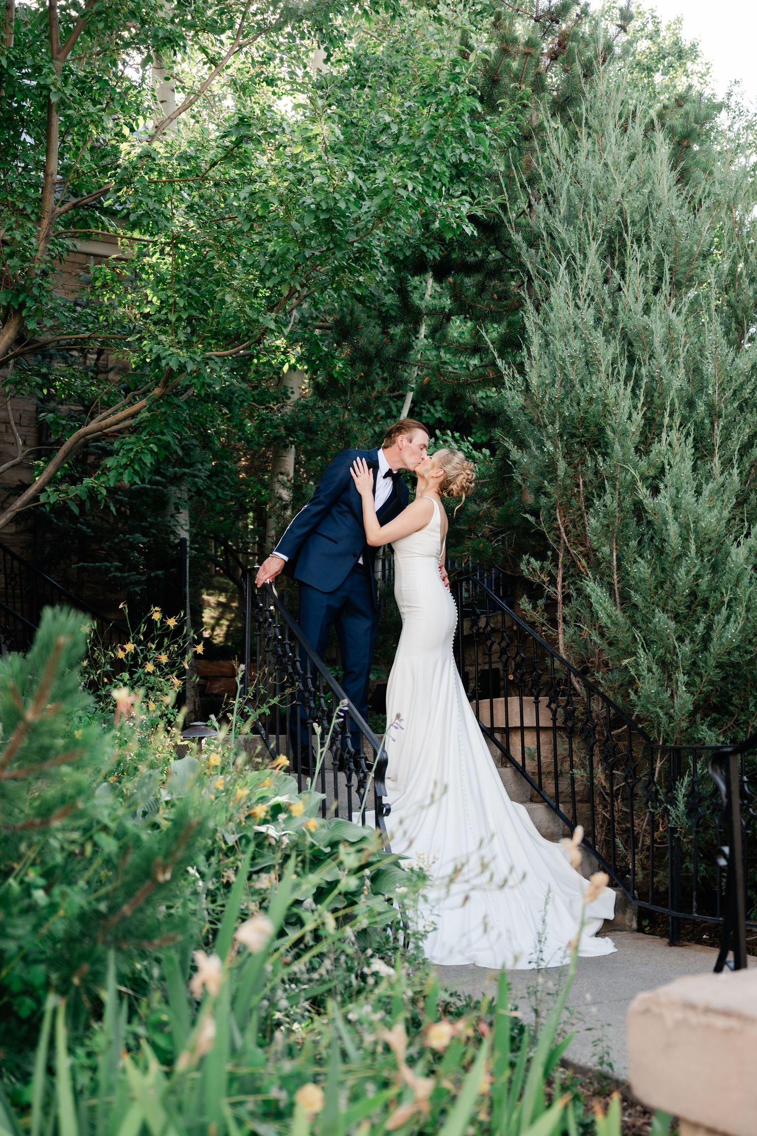 A romantic wedding couple shares an intimate moment on garden steps surrounded by lush greenery.
