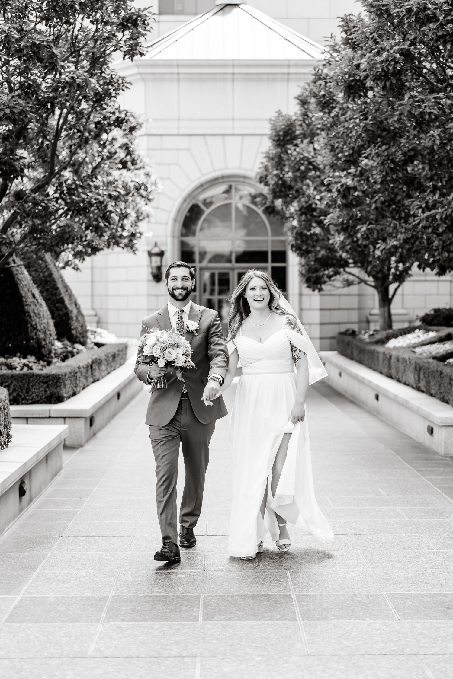 A wedding couple walks joyfully down a stone pathway surrounded by trees and architecture in black and white.