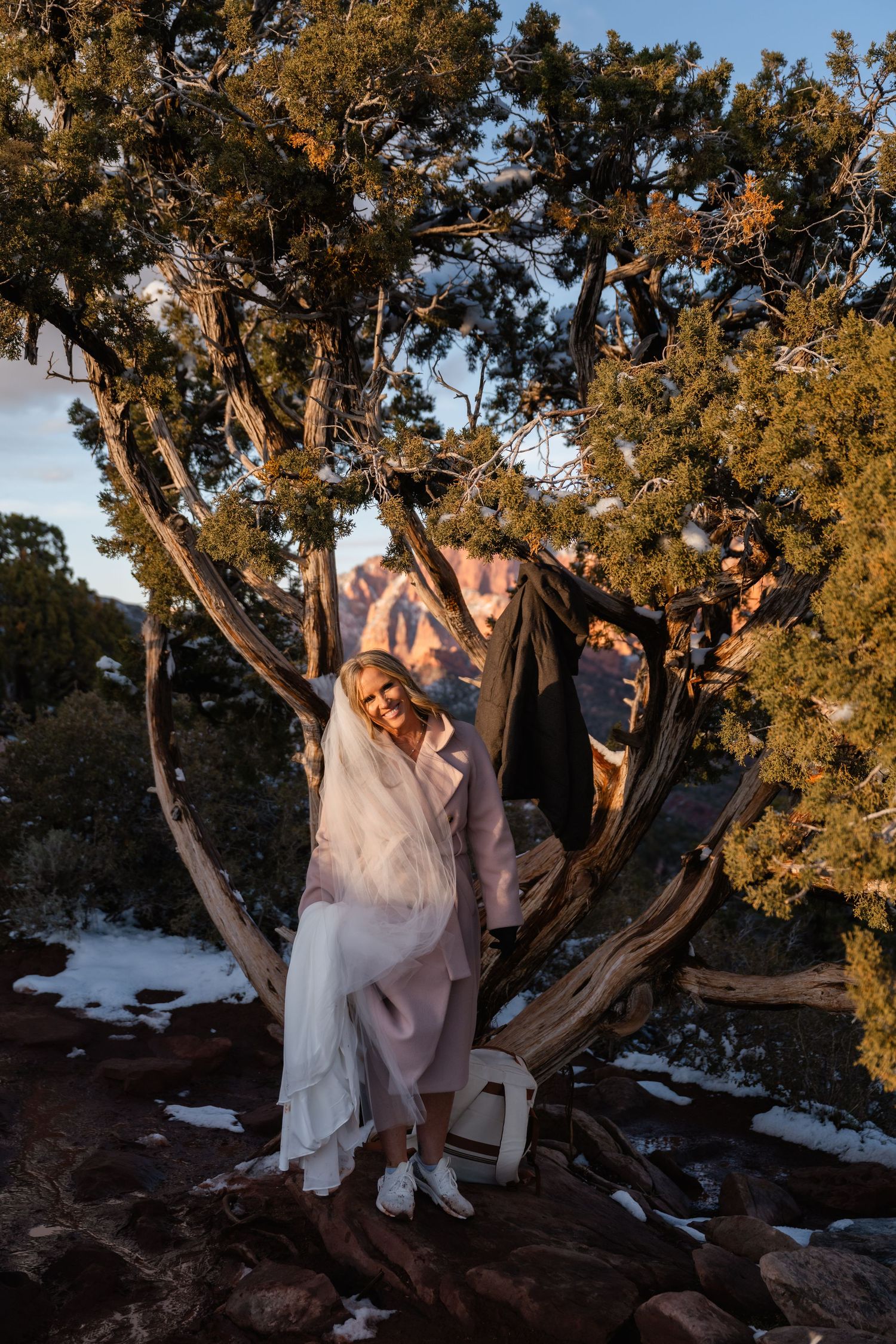 A couple sits playfully among twisted juniper trees on a snowy mountain ledge.
