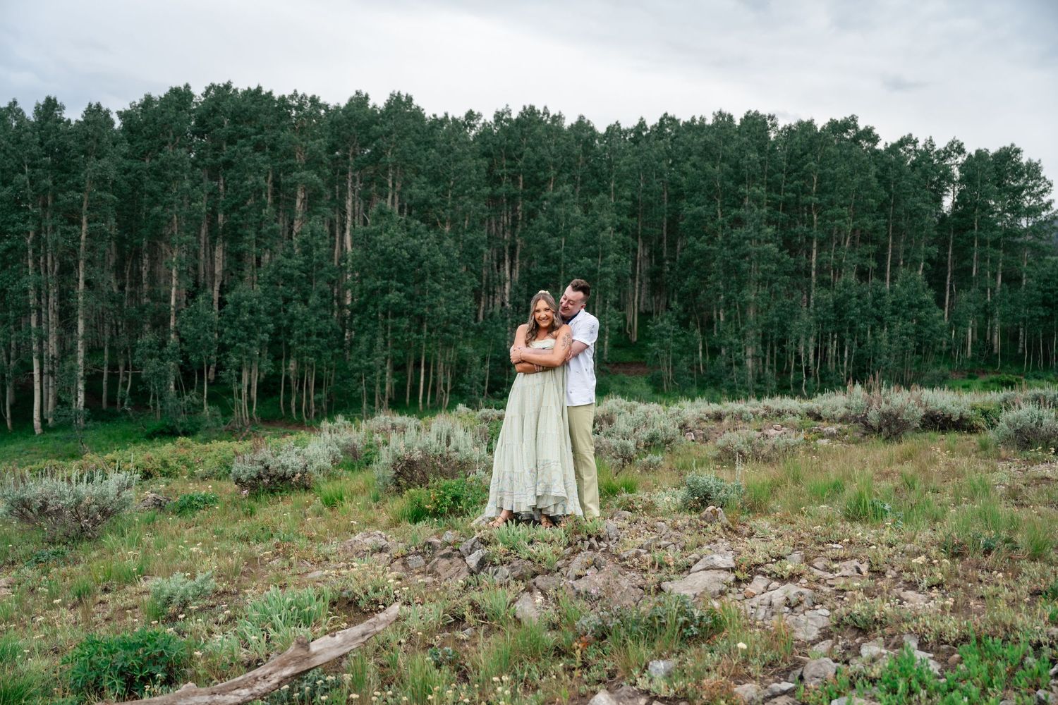 A couple embraces on a hilltop meadow surrounded by tall pine forest in the background.