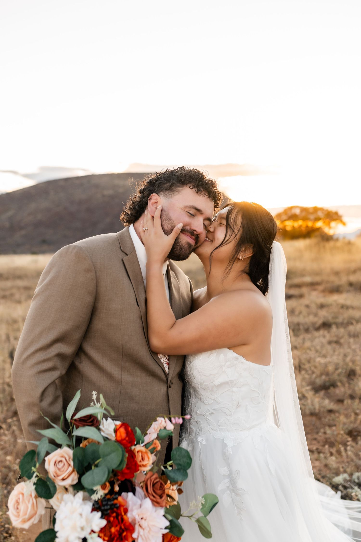 Romantic wedding couple shares intimate moment with autumn-colored bouquet against desert landscape.