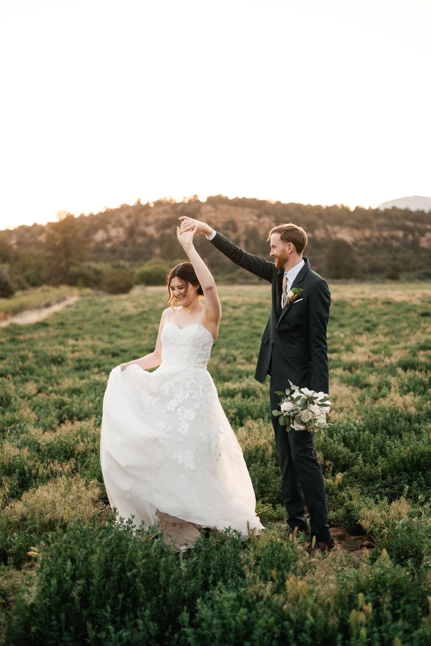 Couple dancing joyfully in a grassy meadow at sunset with flowing white wedding dress.