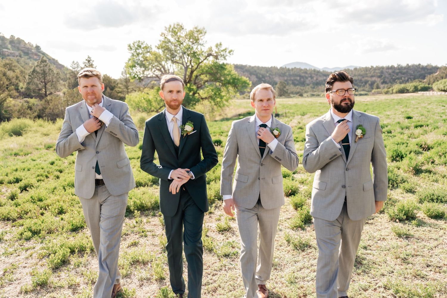 Groomsmen in light gray suits pose playfully with beer bottles in a scenic outdoor setting.