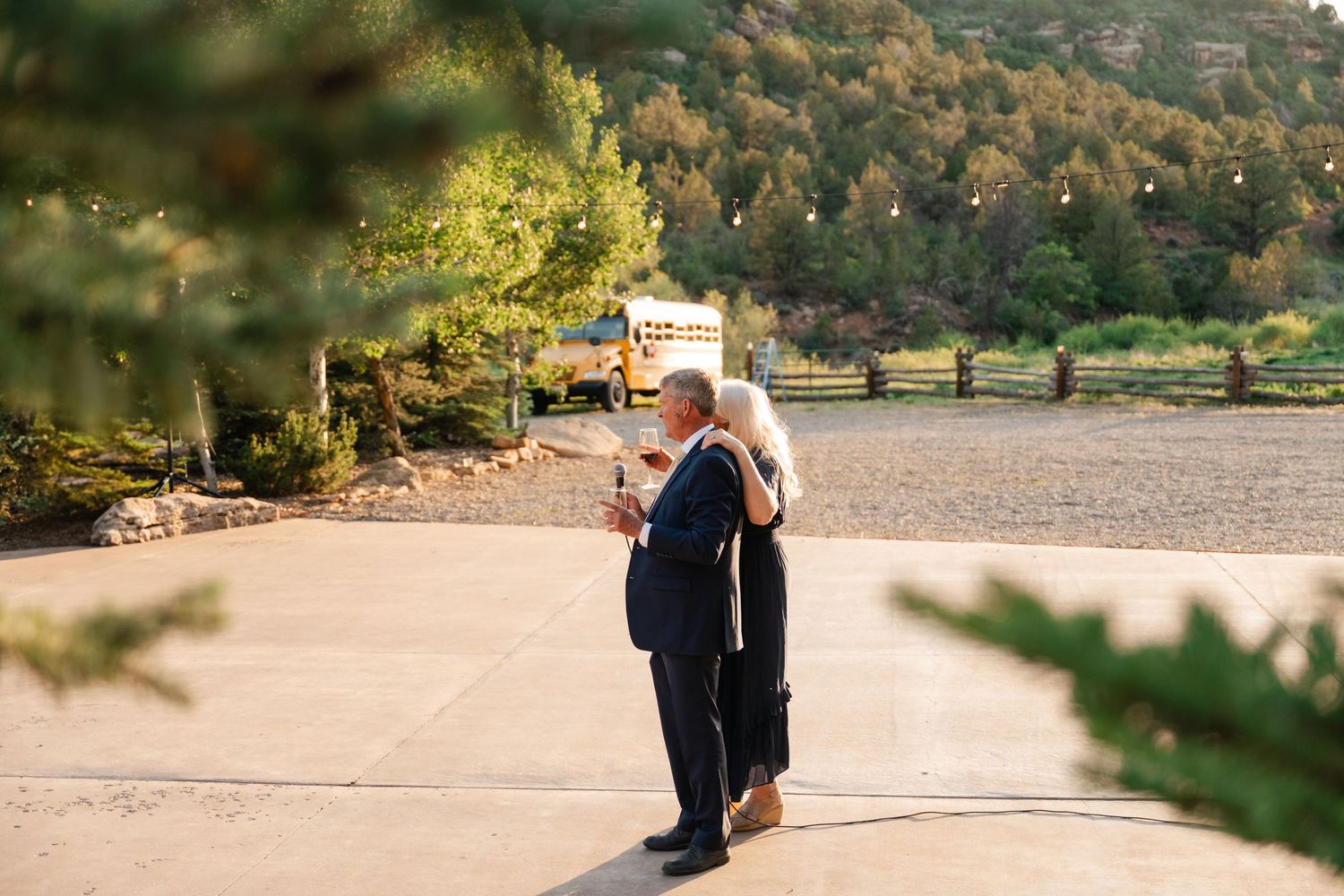 A couple walks along a scenic beach path surrounded by lush greenery and mountains in the background.