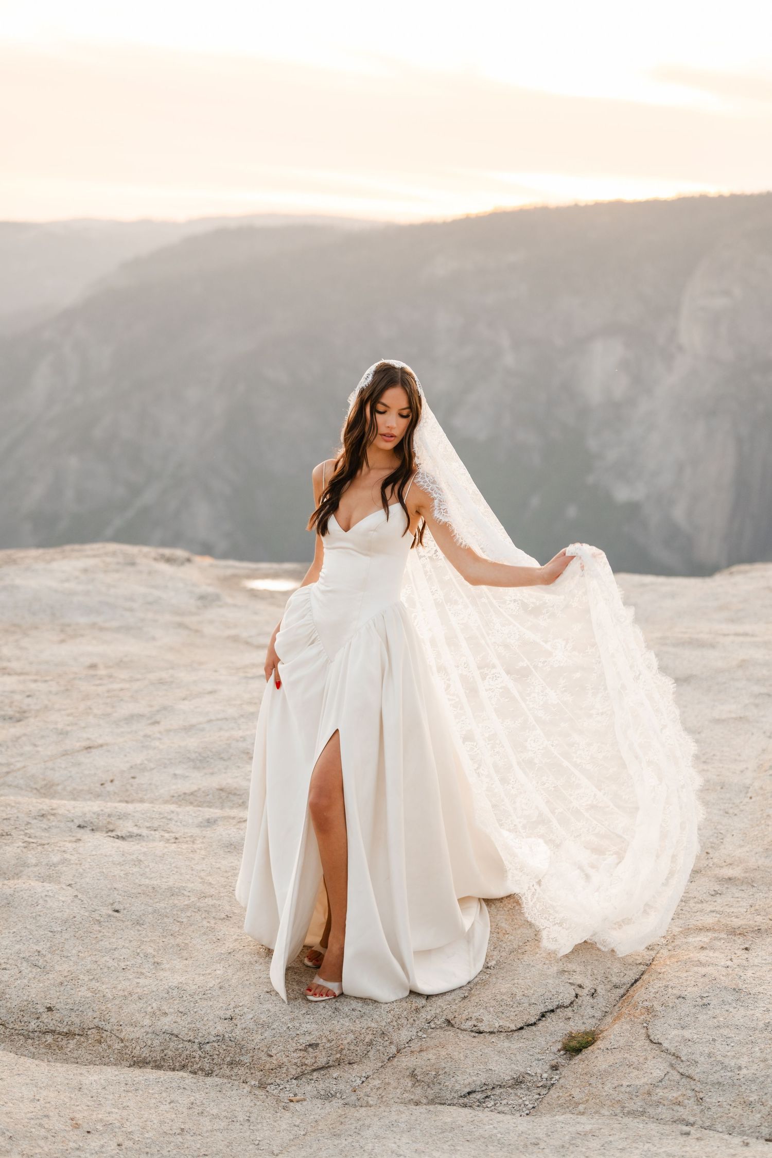 Bride in flowing white gown with lace veil on rocky mountain overlook at golden hour sunset.