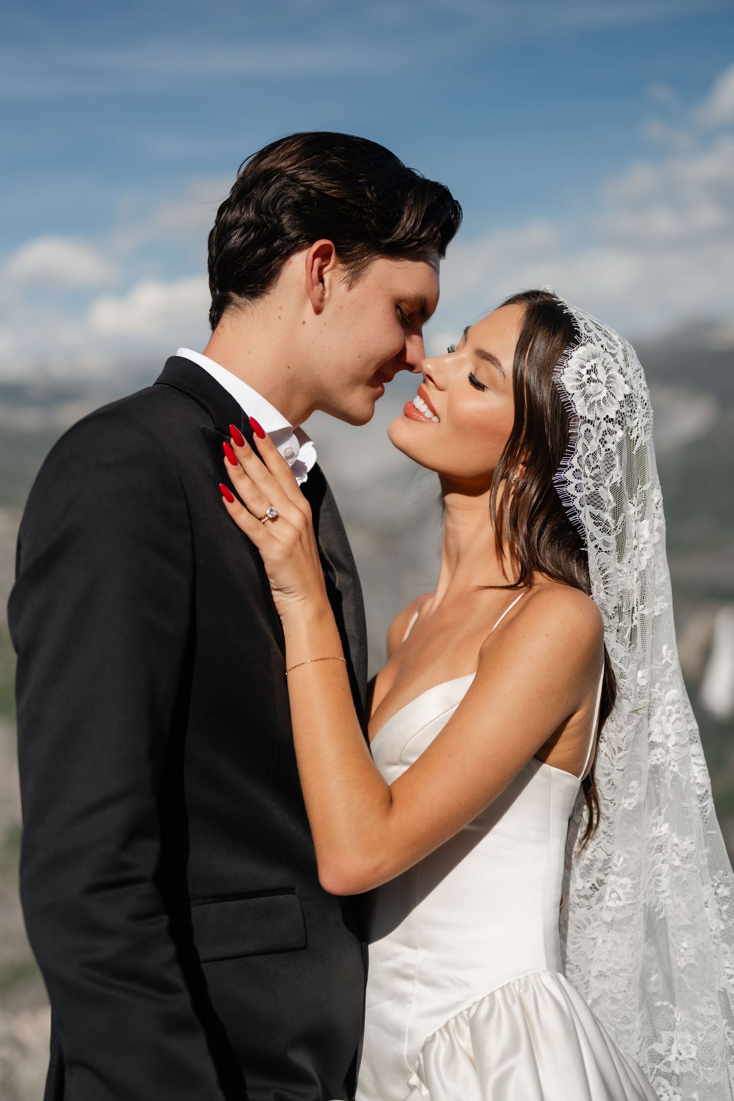 Smiling bride in white dress and lace veil embracing groom in black suit outdoors.