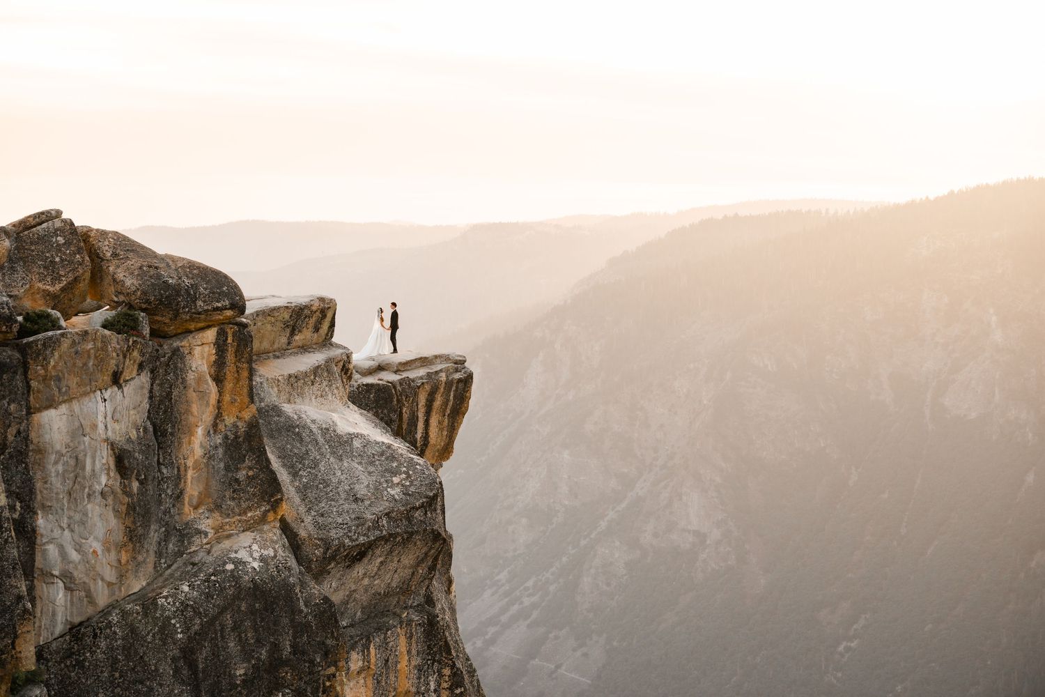 Couple standing on rocky cliff edge at Taft Point at sunset in Yosemite with dramatic valley views below. Yosemite Wedding Photographer.
