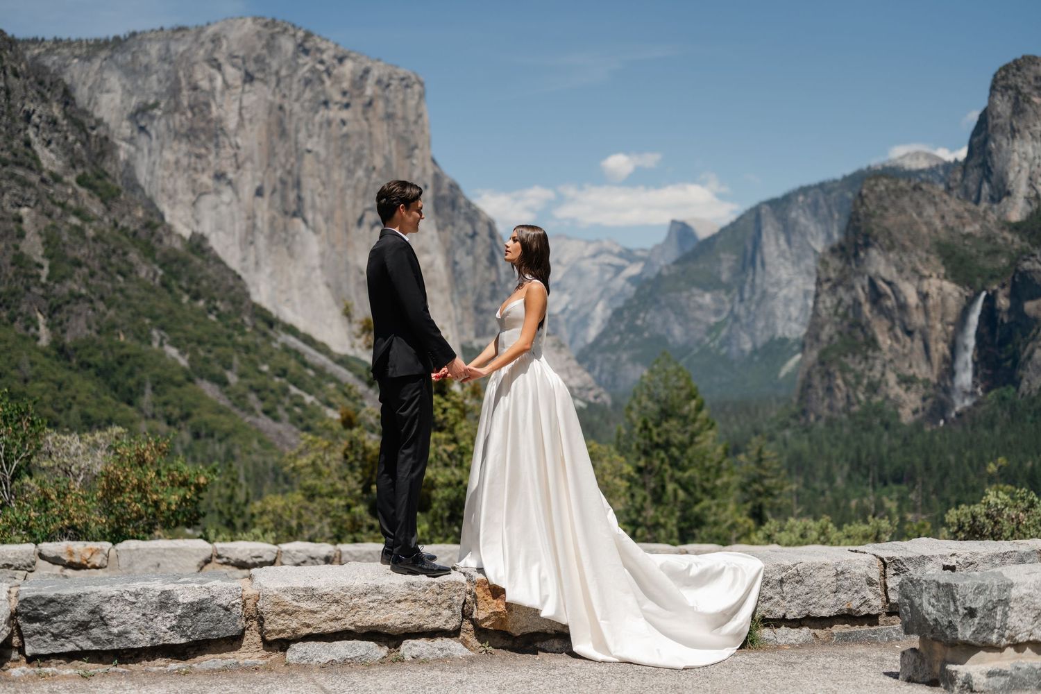 Couple in wedding attire holding hands at Yosemite Valley overlook with Half Dome and granite cliffs backdrop.
