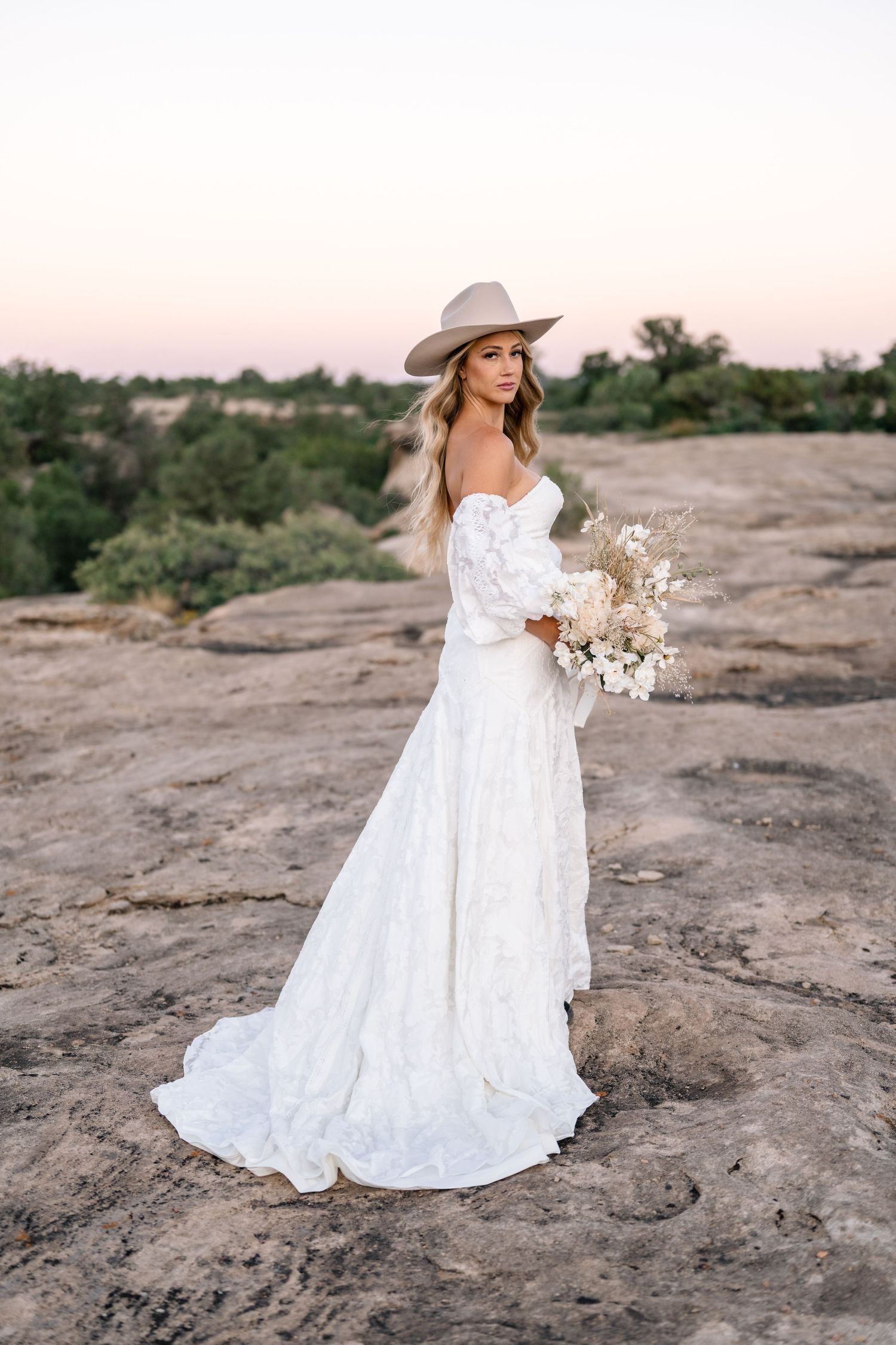 Elegant bridal portrait featuring western style dress and hat against desert landscape at sunset.
