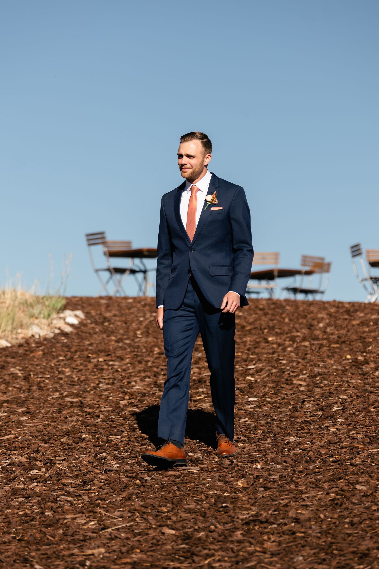 A person in a navy suit and orange tie stands on brown soil with outdoor seating in the background. Park City Wedding Photographer.