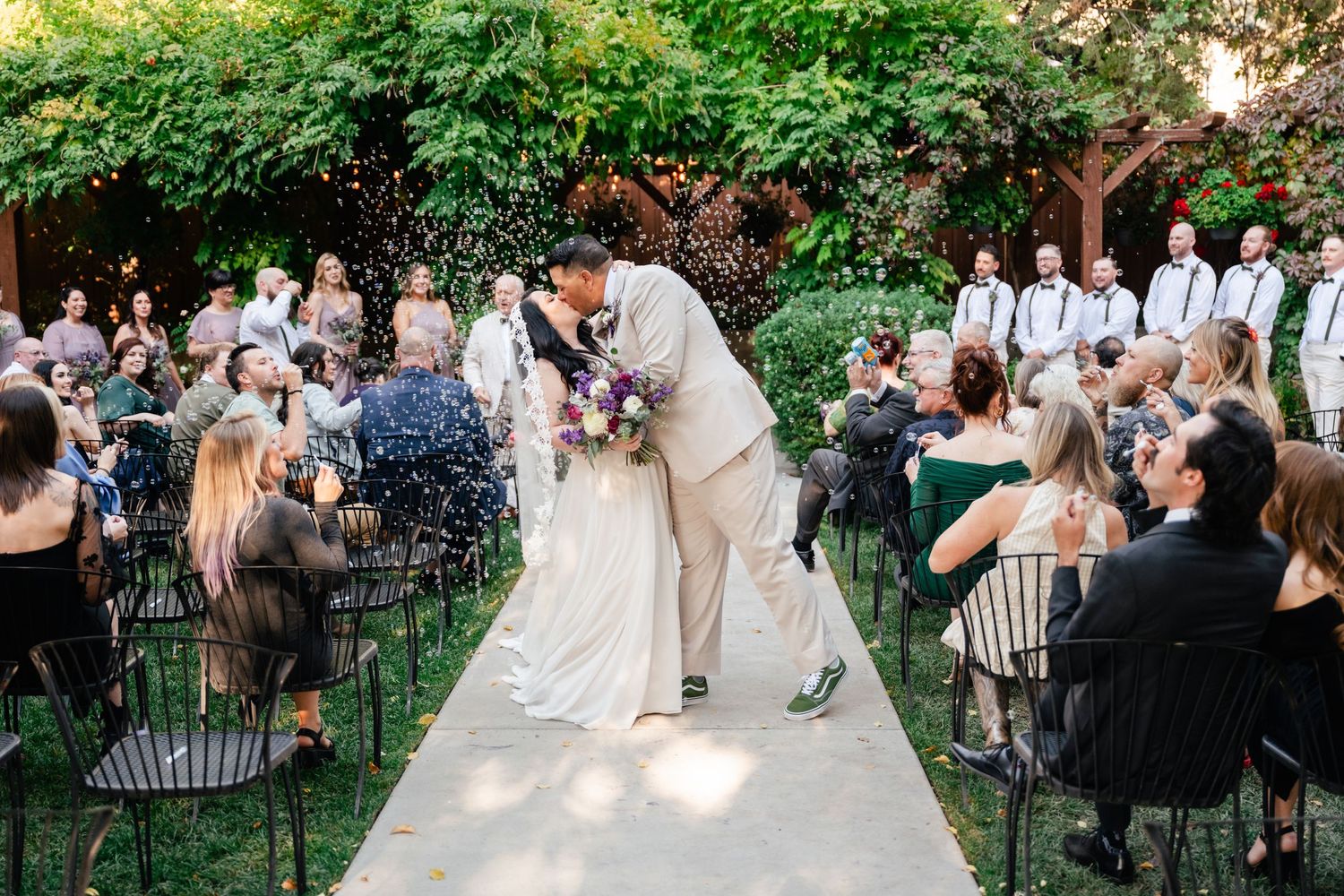 A romantic moment between newlyweds on a garden path surrounded by guests at an outdoor summer wedding ceremony.