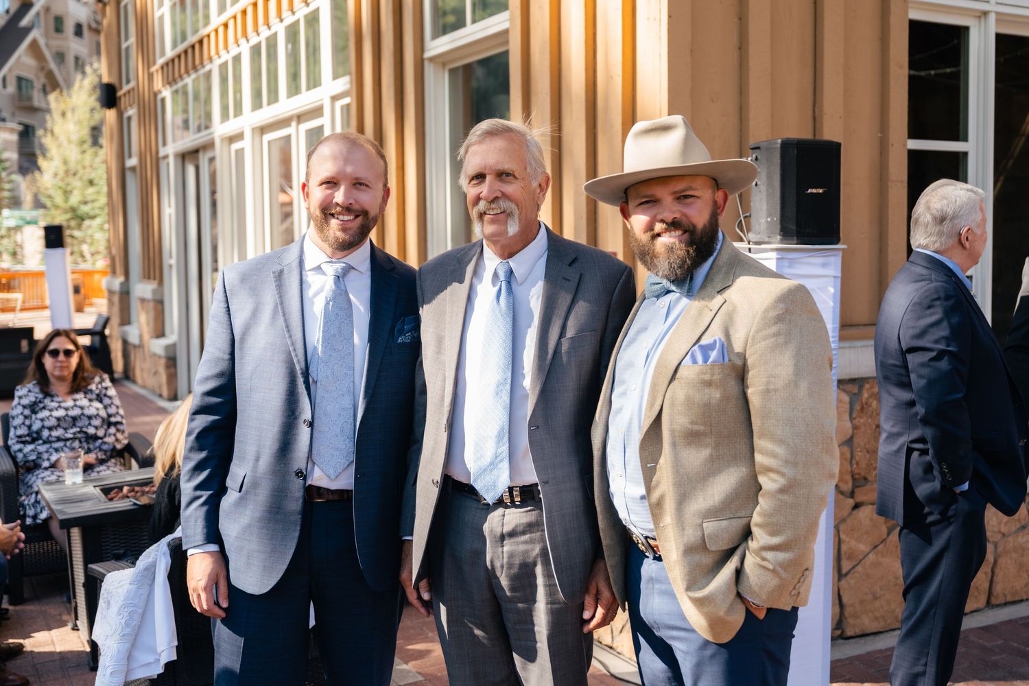 Three men dressed in business attire pose together and smile outside a building with classic architecture. Park City Wedding Photographer.