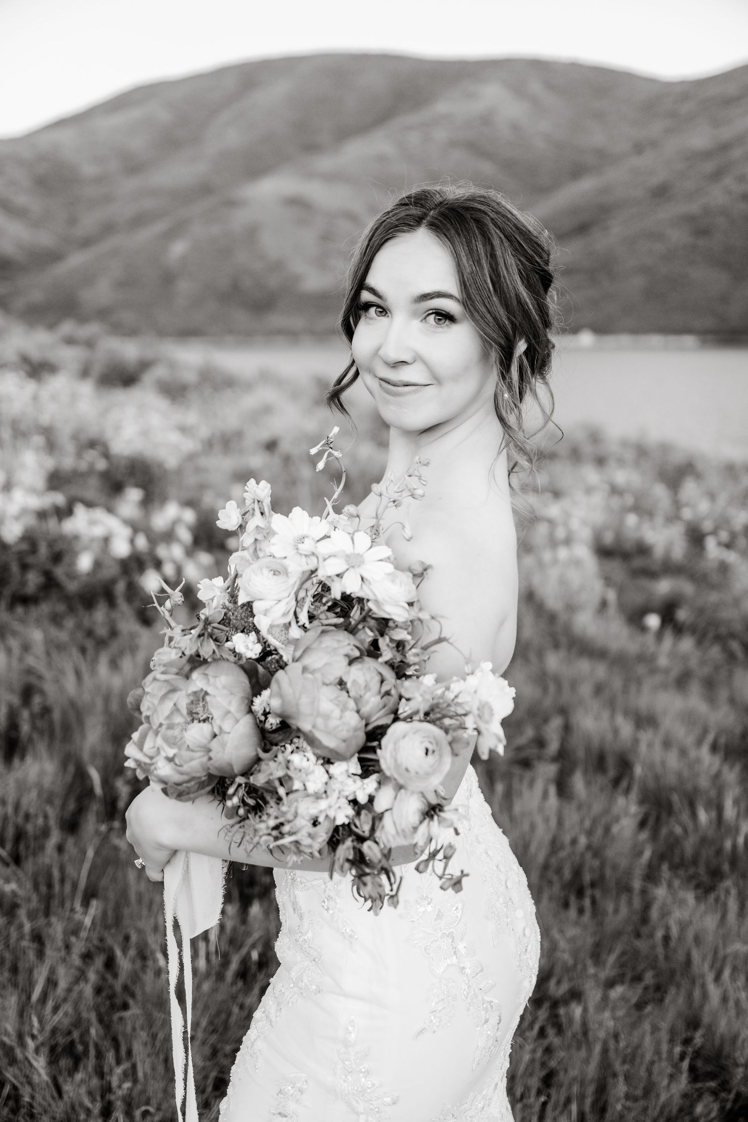 Bride in lace dress poses with elegant peony bouquet against mountainous landscape in black and white.