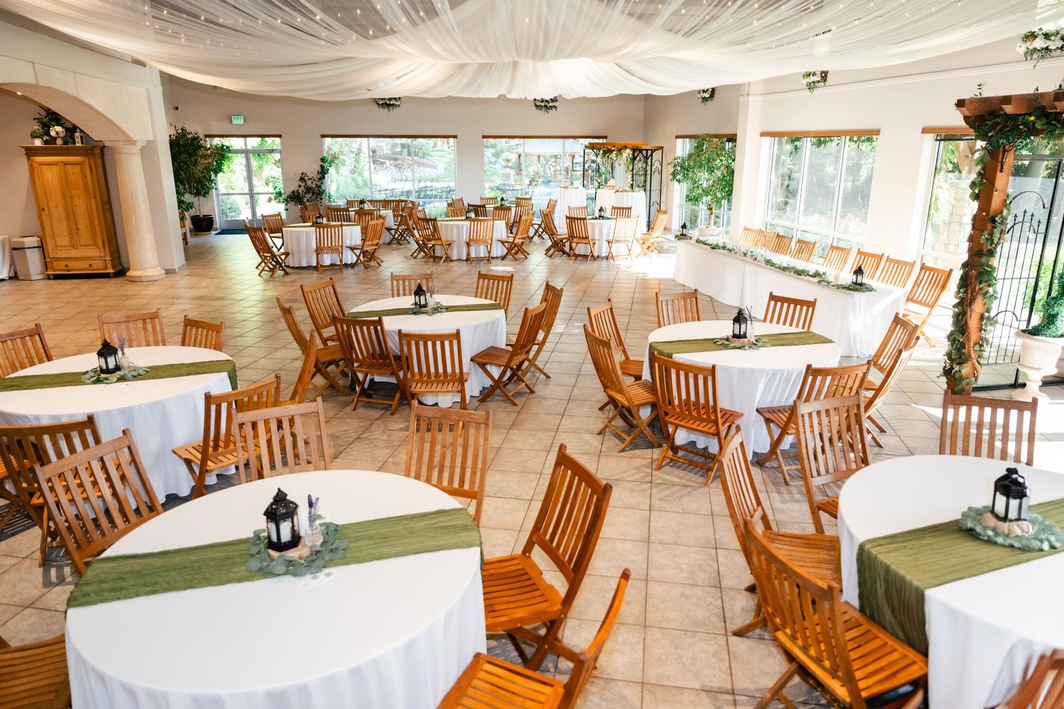Elegant indoor dining area with white tablecloths, wooden chairs and sheer fabric draped from ceiling in a wedding reception setup.