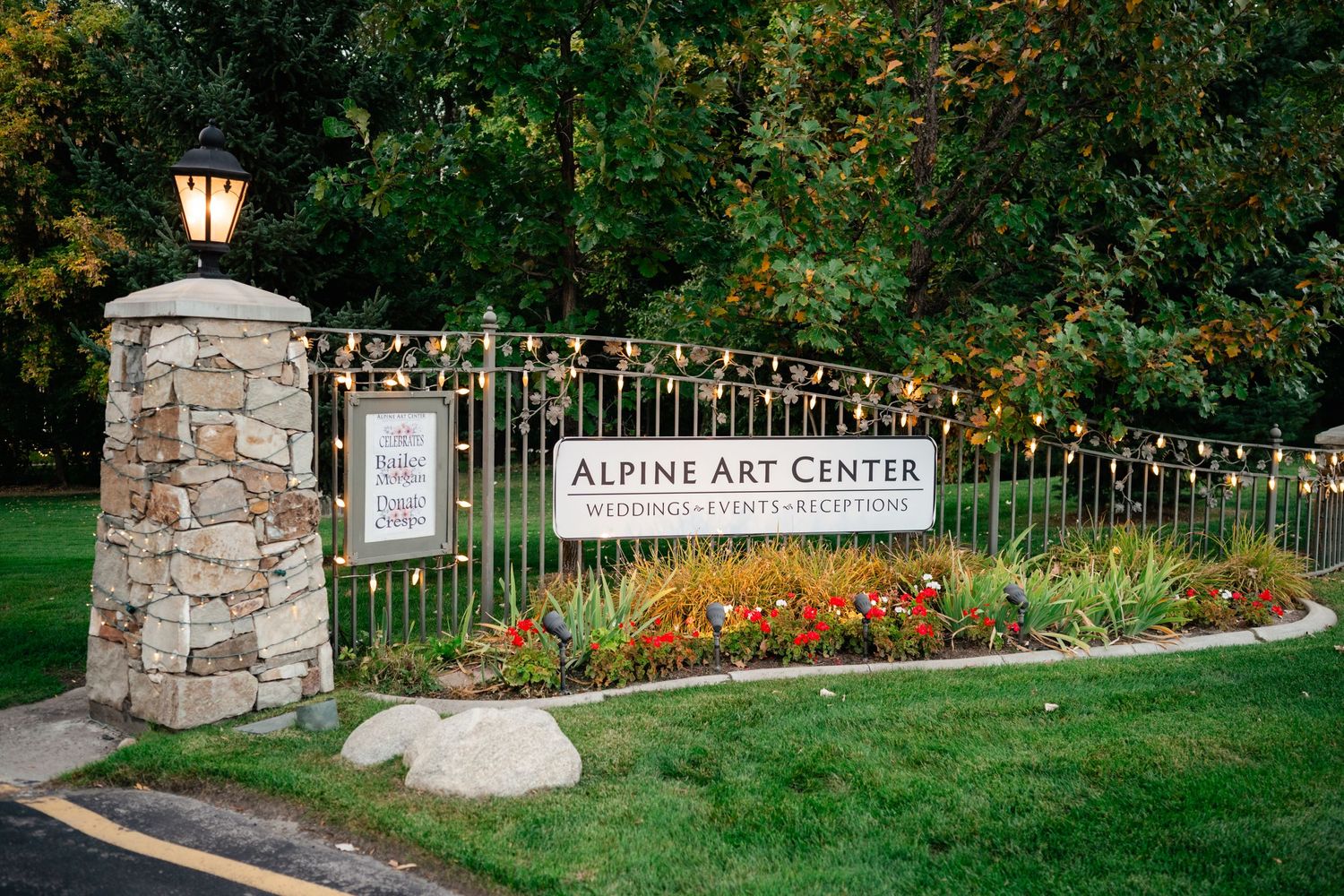 Stone entrance pillar with sign reading Alpine Art Centre surrounded by colorful flowers and string lights in garden setting.