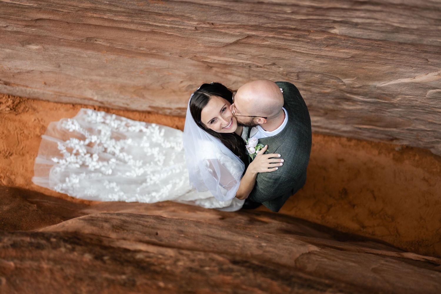 A couple in wedding attire embrace while standing on rustic wooden flooring viewed from above.