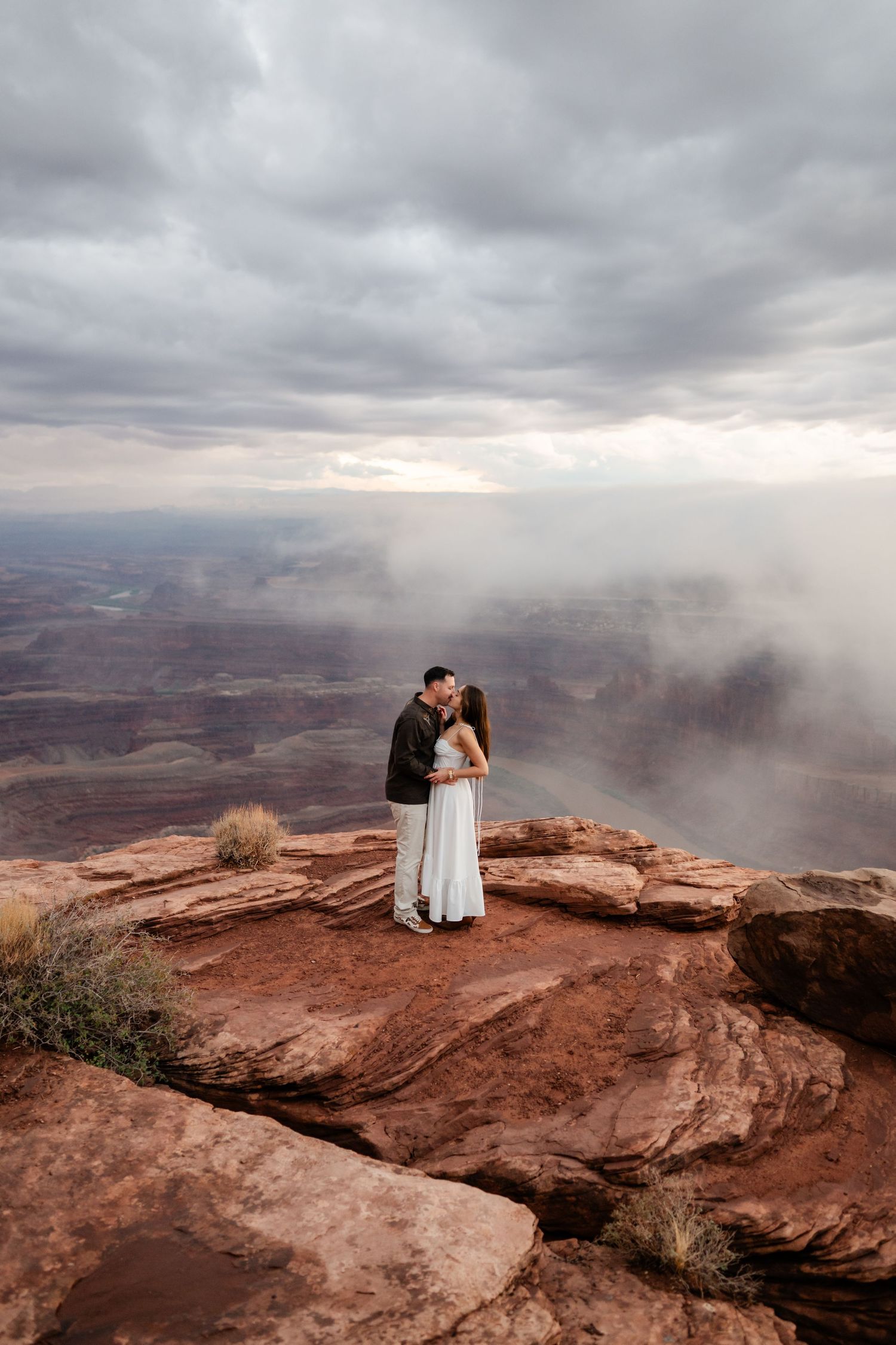 A couple embraces on red rock cliffs overlooking a dramatic desert canyon landscape under moody storm clouds.