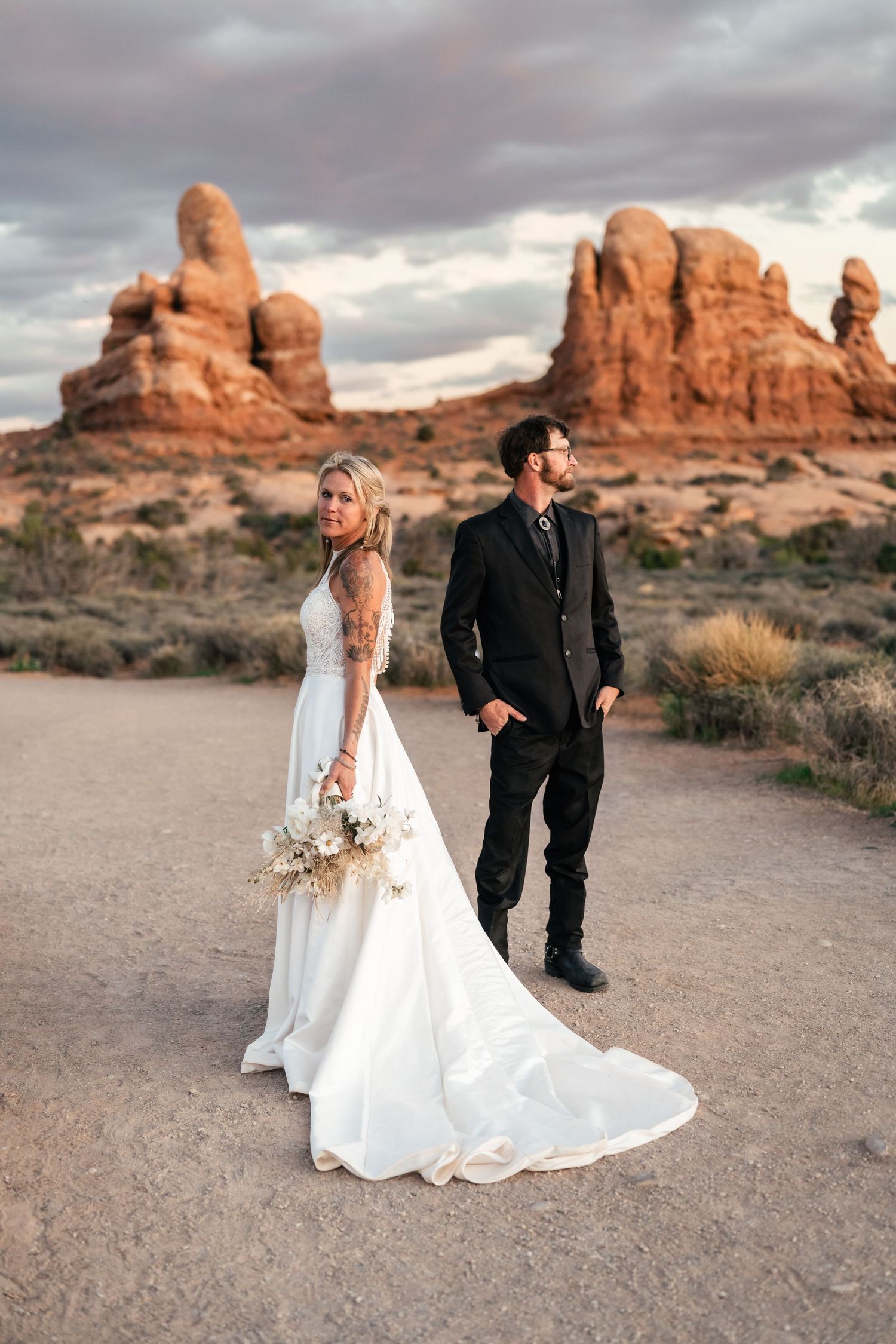 Wedding couple stands against backdrop of majestic red rock formations at sunset in desert landscape.