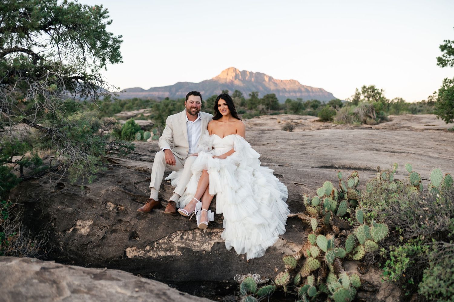 Zion Adventure Session. A couple in wedding attire sits on red rocks with desert landscape and mountains in the background at sunset.