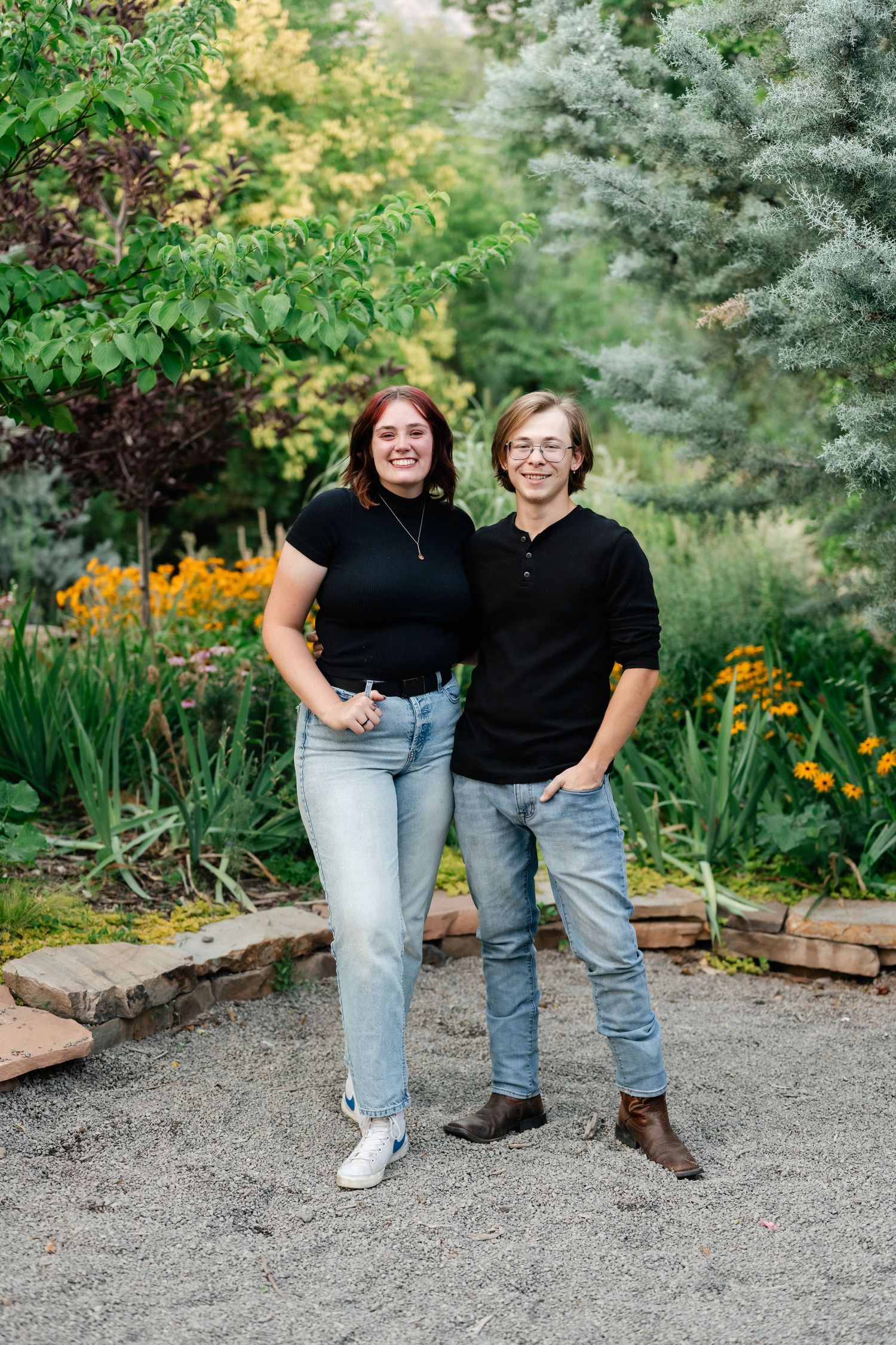 Two people in black tops and jeans pose casually together in a garden setting with colorful flowers and evergreens.