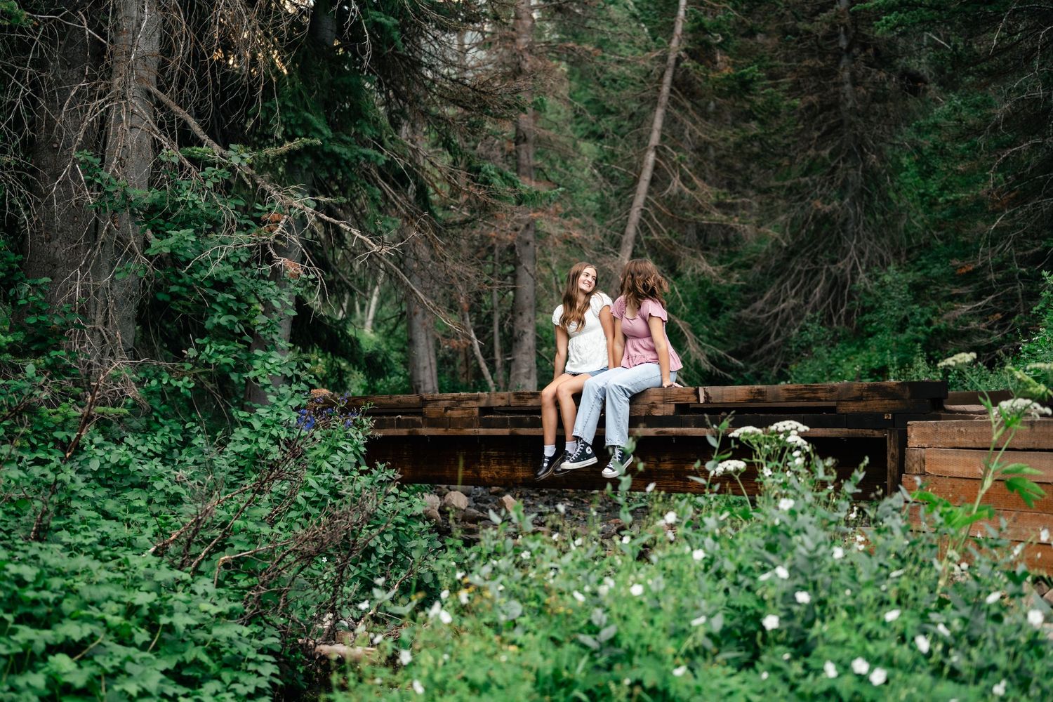 A couple sits on a rustic wooden bench surrounded by lush forest greenery and blooming wildflowers.