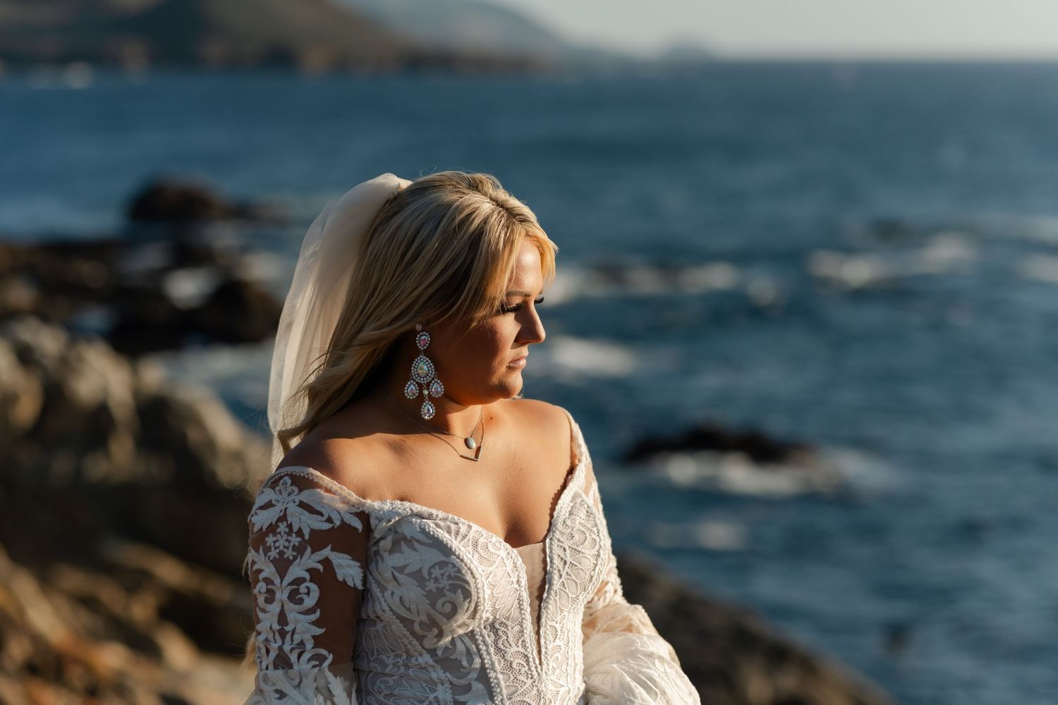 Person in lace wedding dress poses against rocky ocean coastline at sunset.