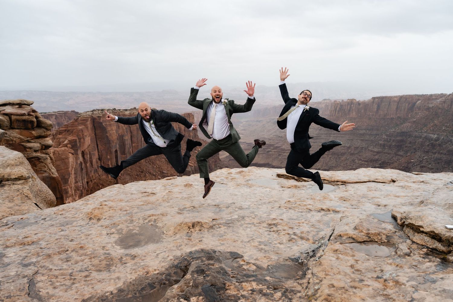 Wedding party members jump joyfully on desert cliff edge with canyon backdrop.