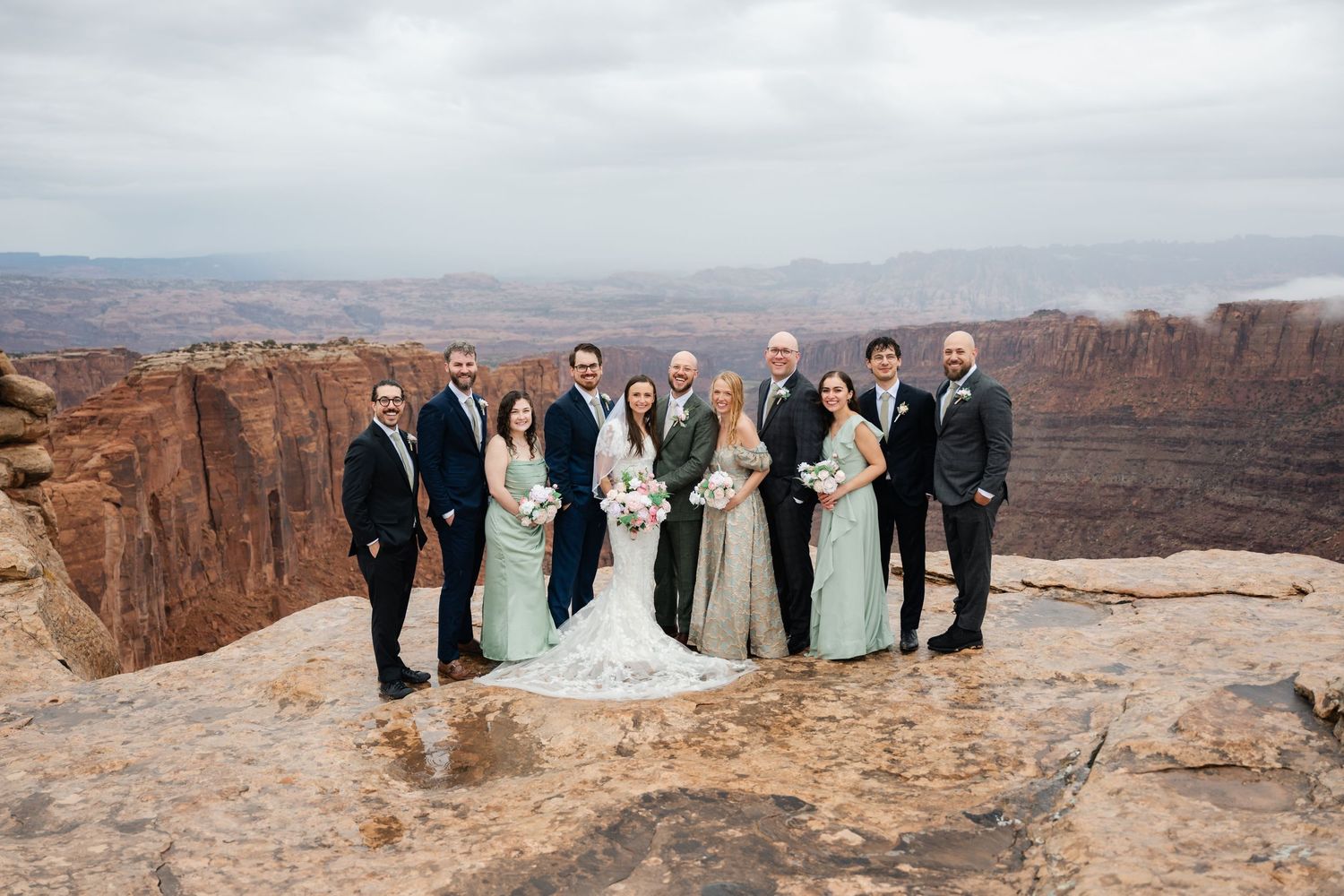 A wedding party poses for photos on the edge of dramatic red rock cliffs at sunset in a desert landscape.