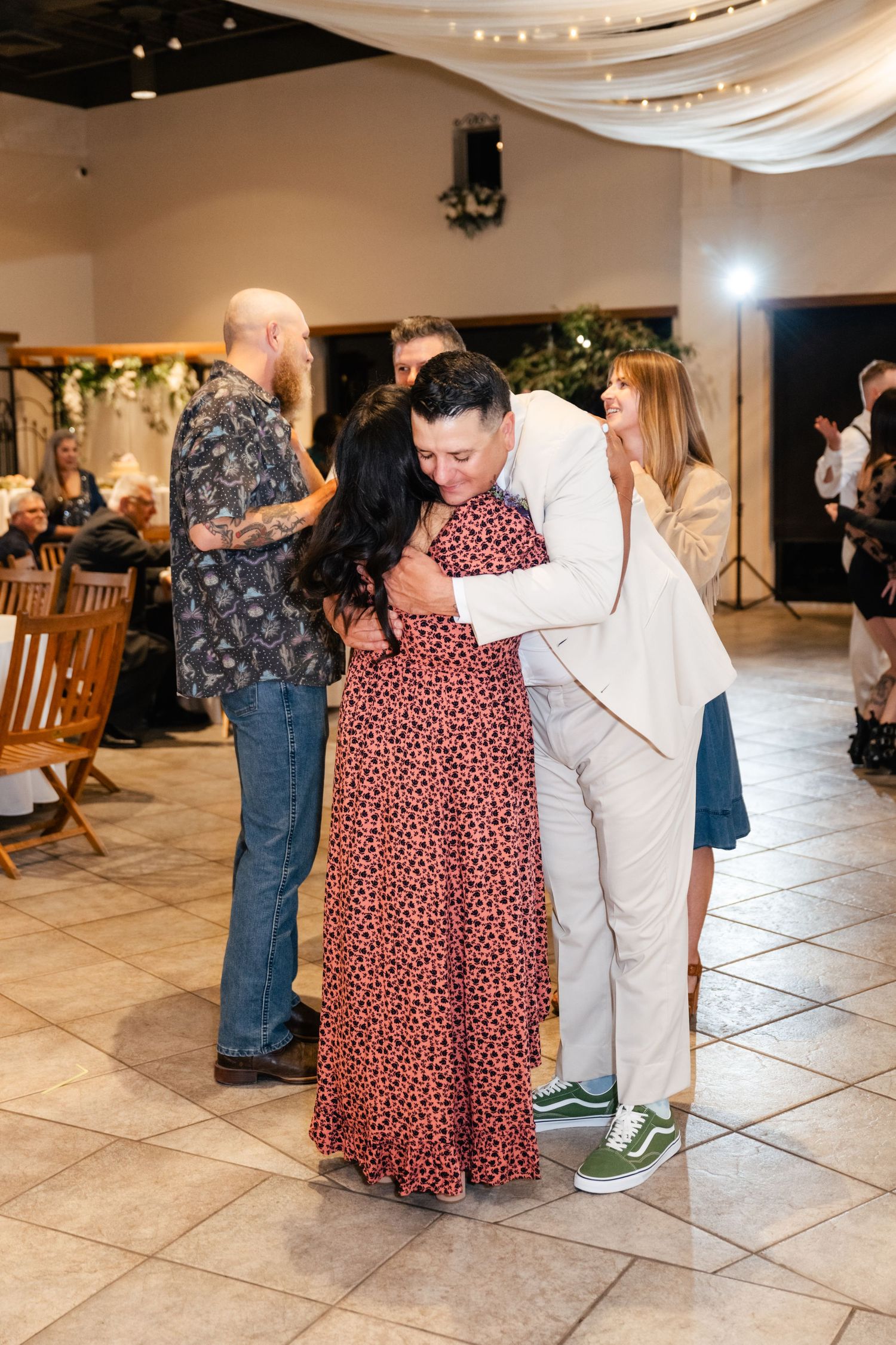 Wedding guests share an emotional embrace on a tiled dance floor decorated with white fabric and lights.