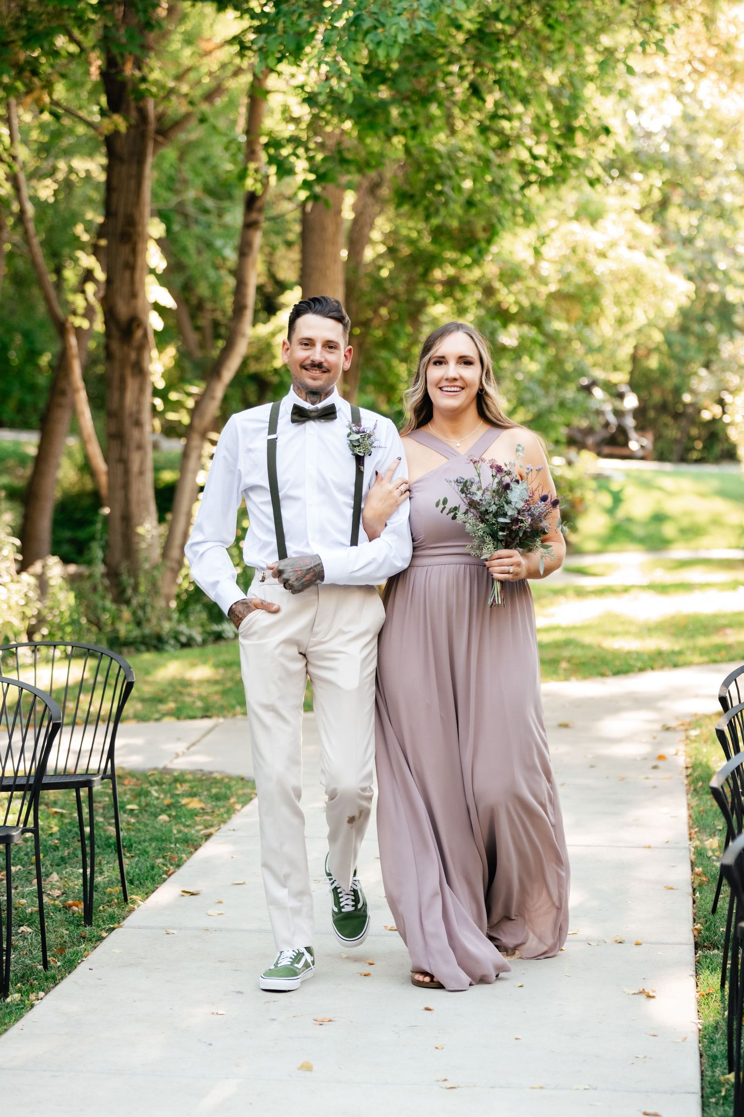 Wedding attendants walk down garden path, with groomsman in white suit and bridesmaid in flowing mauve dress.